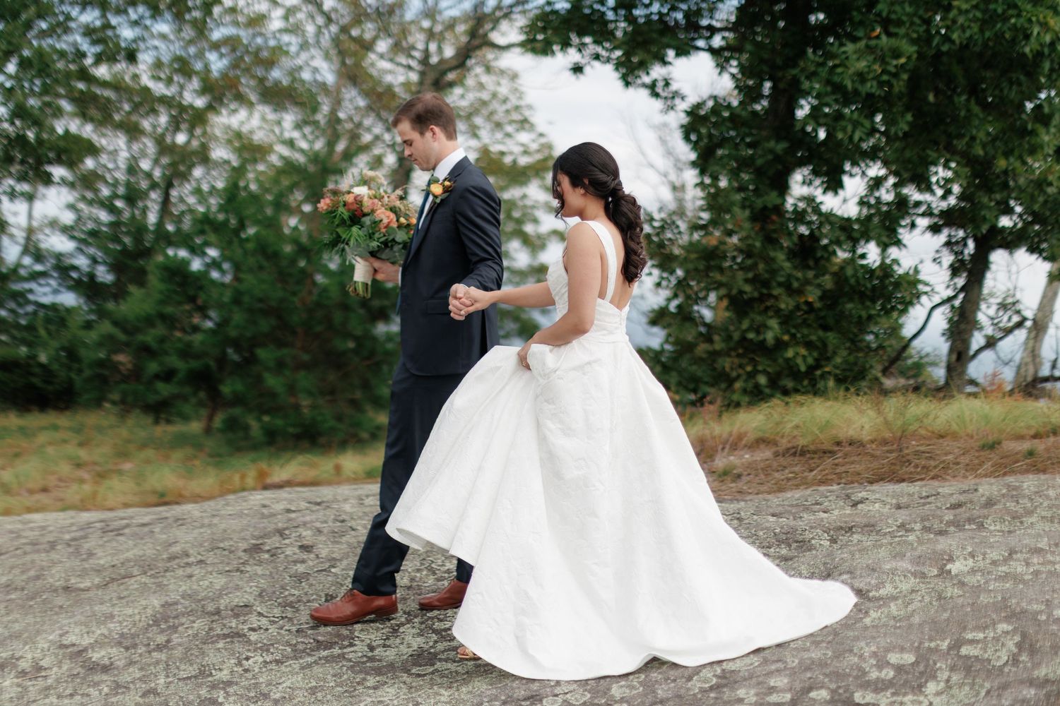 Bride in white gown and groom in navy suit walk hand-in-hand on stone path with trees in background.