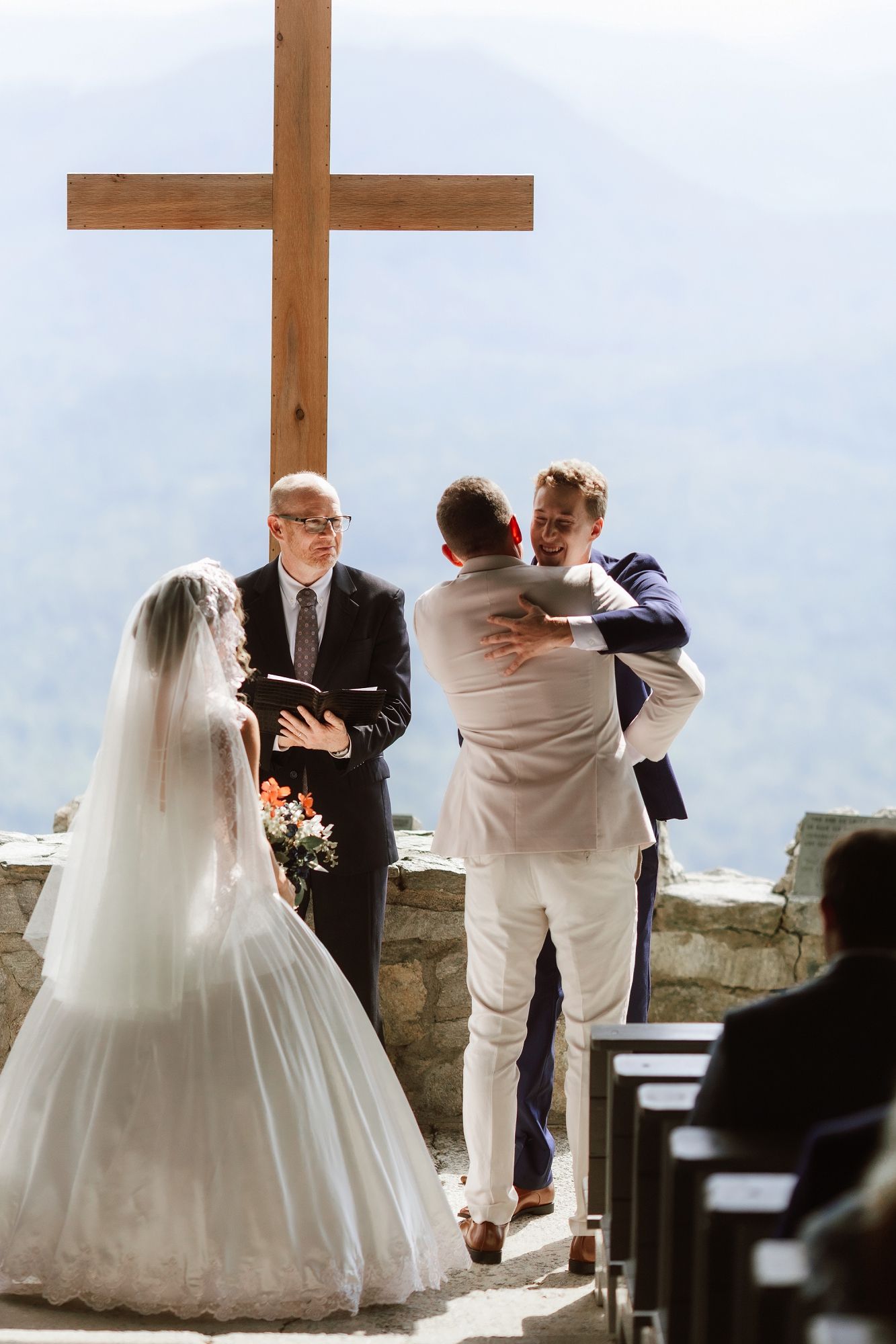 Outdoor wedding ceremony at a mountain chapel with a wooden cross, bride in white gown, groom embracing best man.