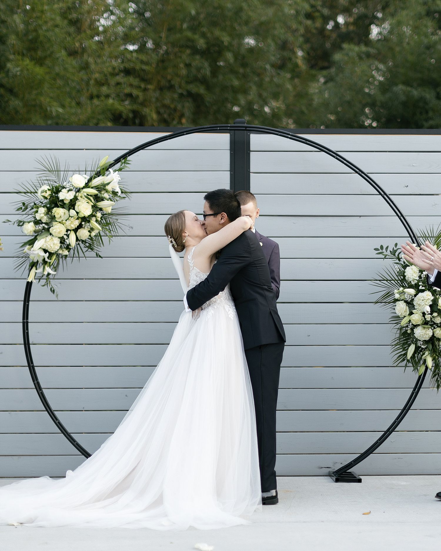 Newlyweds share their first kiss under a circular arch decorated with white flowers against a gray wall.