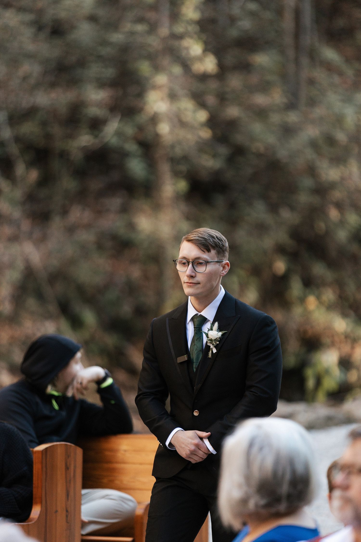 A groom in a black suit and glasses stands at an outdoor wedding ceremony with guests seated nearby.