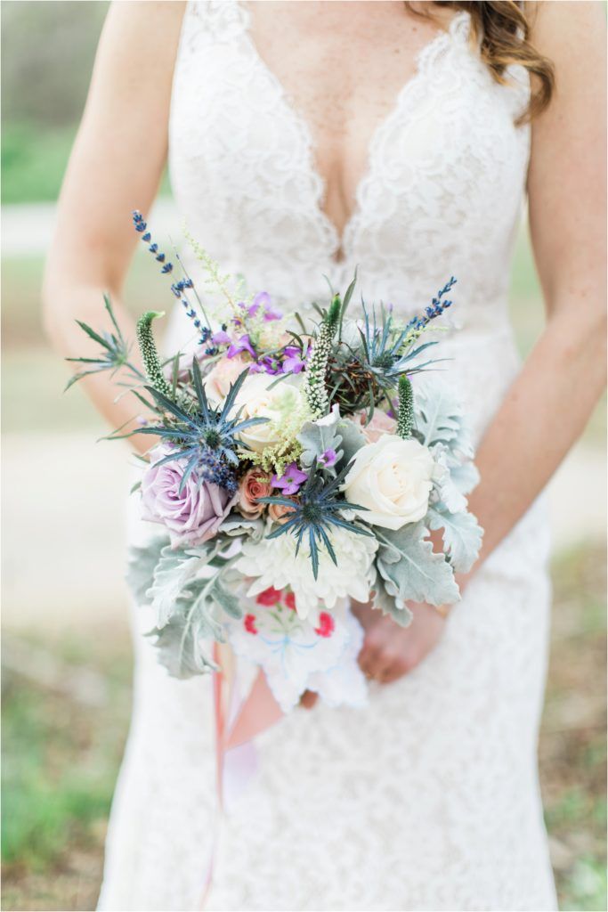 A delicate bridal bouquet featuring white roses, lavender, and blue thistle with a lace wedding dress.