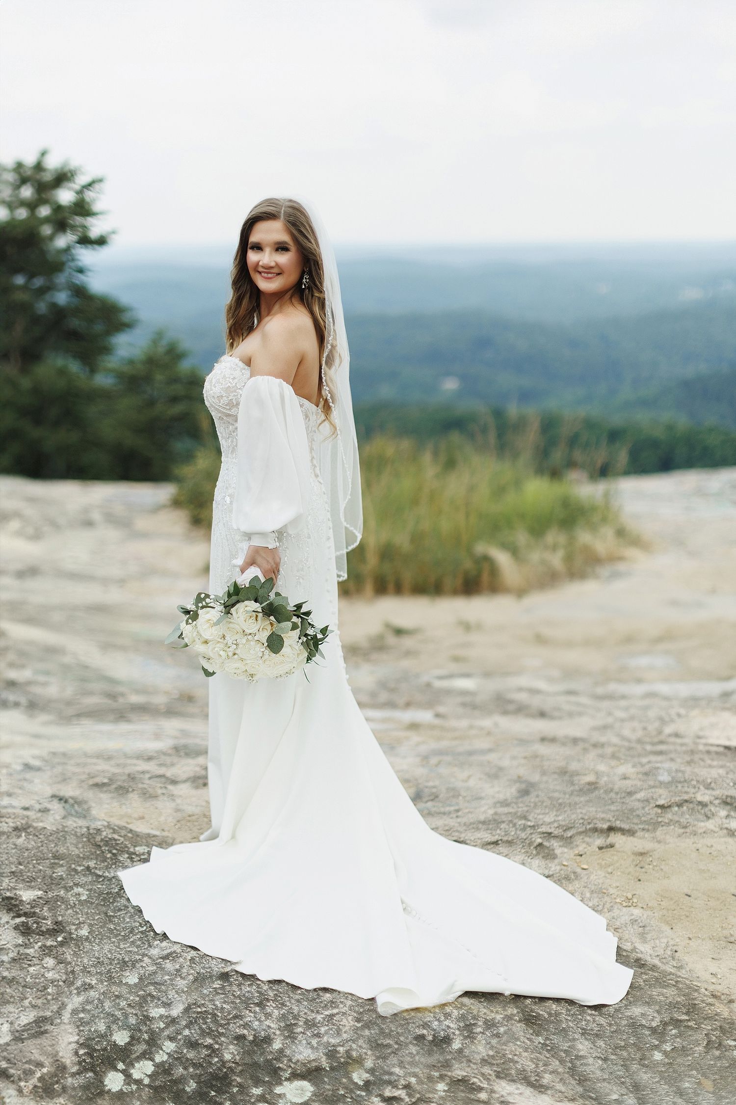 A bride in a white off-shoulder gown poses with bouquet against mountain vista backdrop.