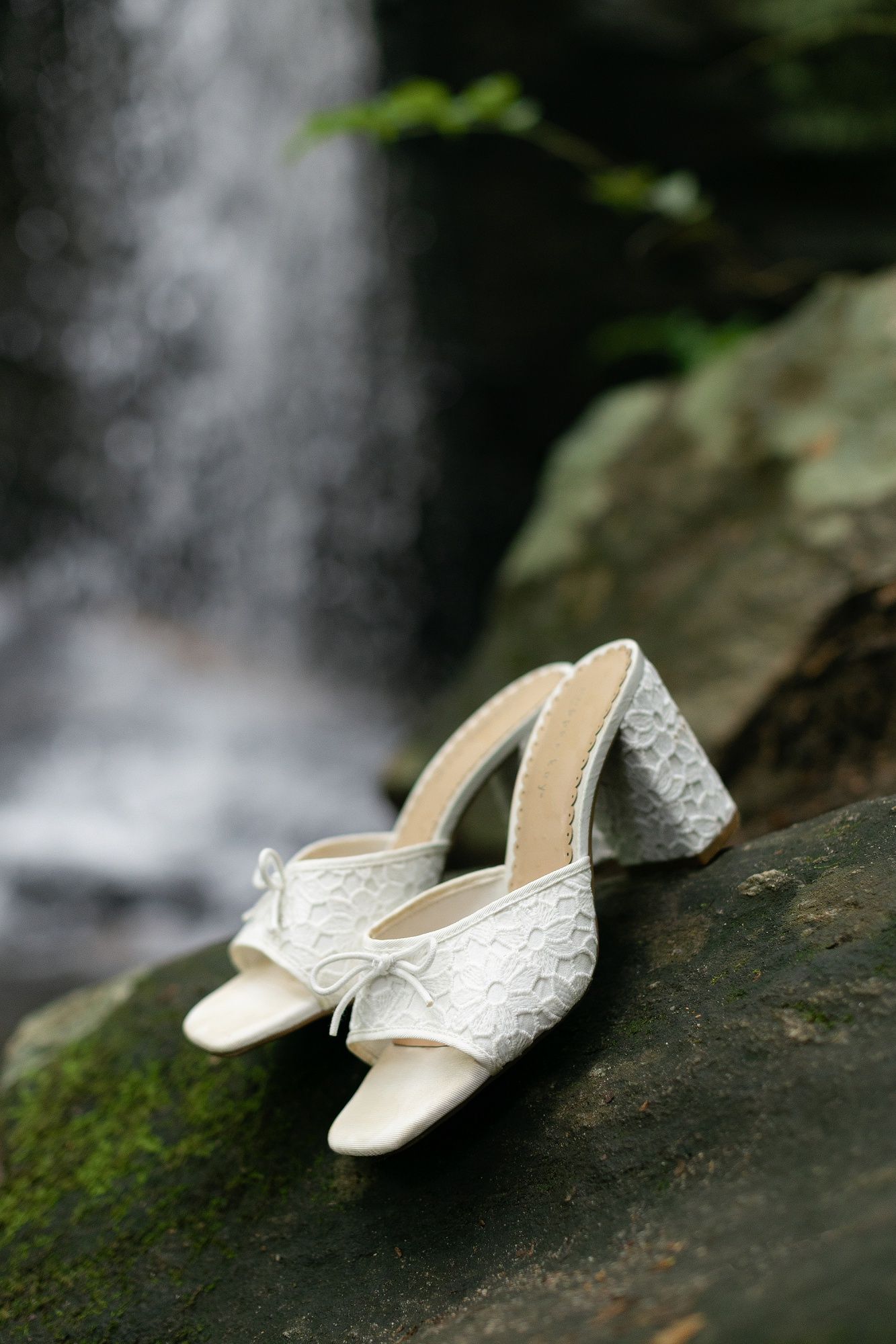 Elegant white lace wedding shoes with low heels placed on mossy rock near a cascading waterfall.