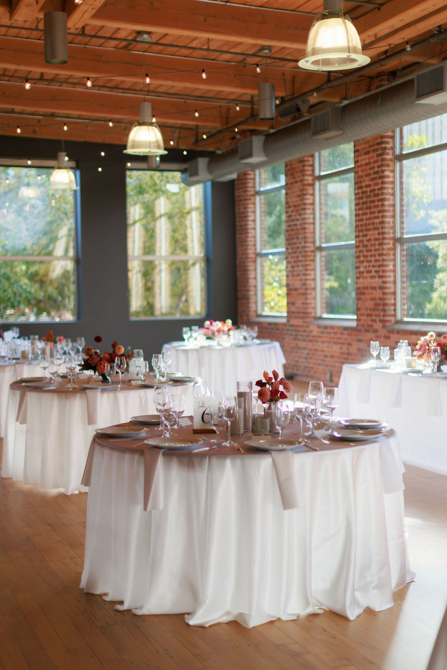 Elegant wedding reception hall with white draped round tables, red brick walls, hardwood floors, and string lights overhead.