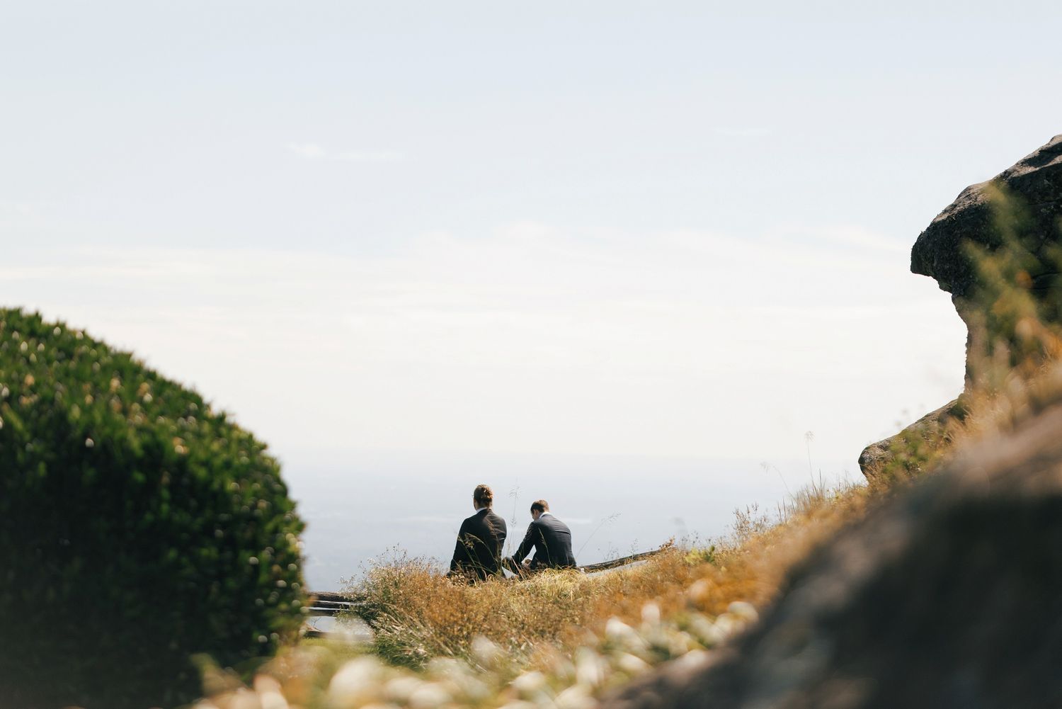 Two silhouettes sit together on a hilltop overlooking a scenic mountain landscape at sunset.
