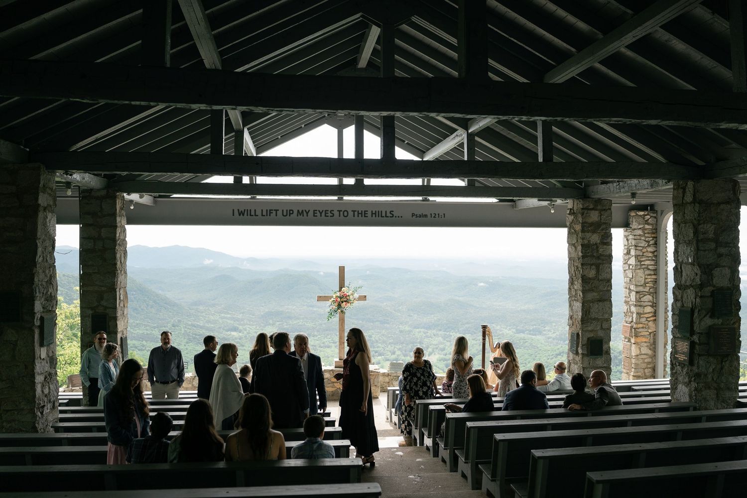 A group of people gather at a mountaintop open-air chapel with a wooden cross and stunning valley views through stone pillars.