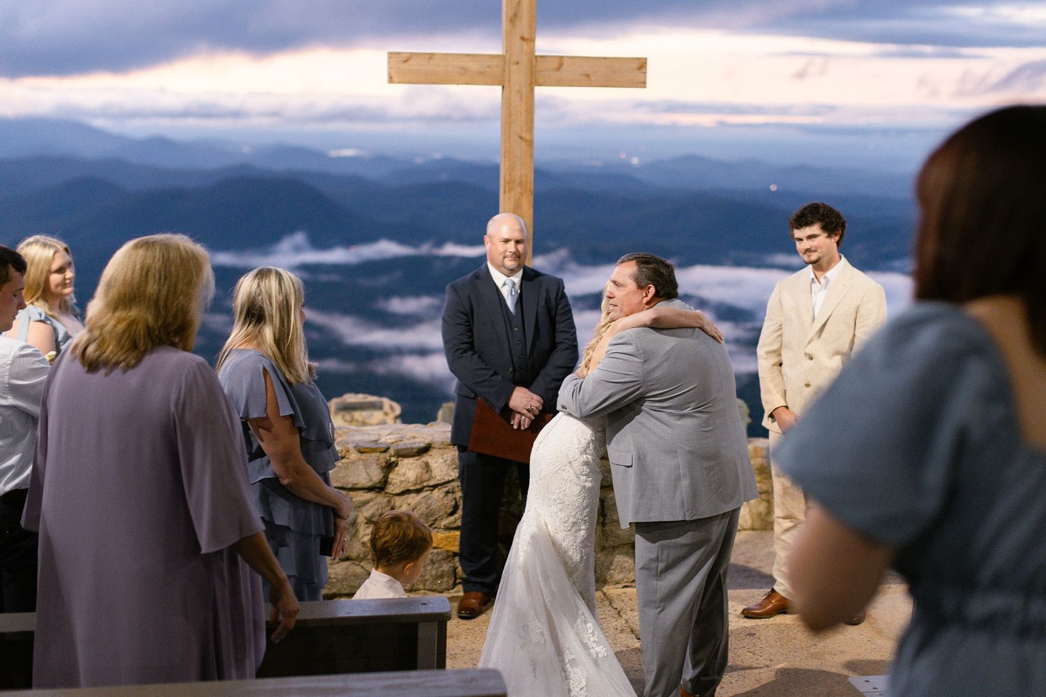 A mountain wedding ceremony takes place at scenic overlook with a wooden cross and guests gathered at sunset.