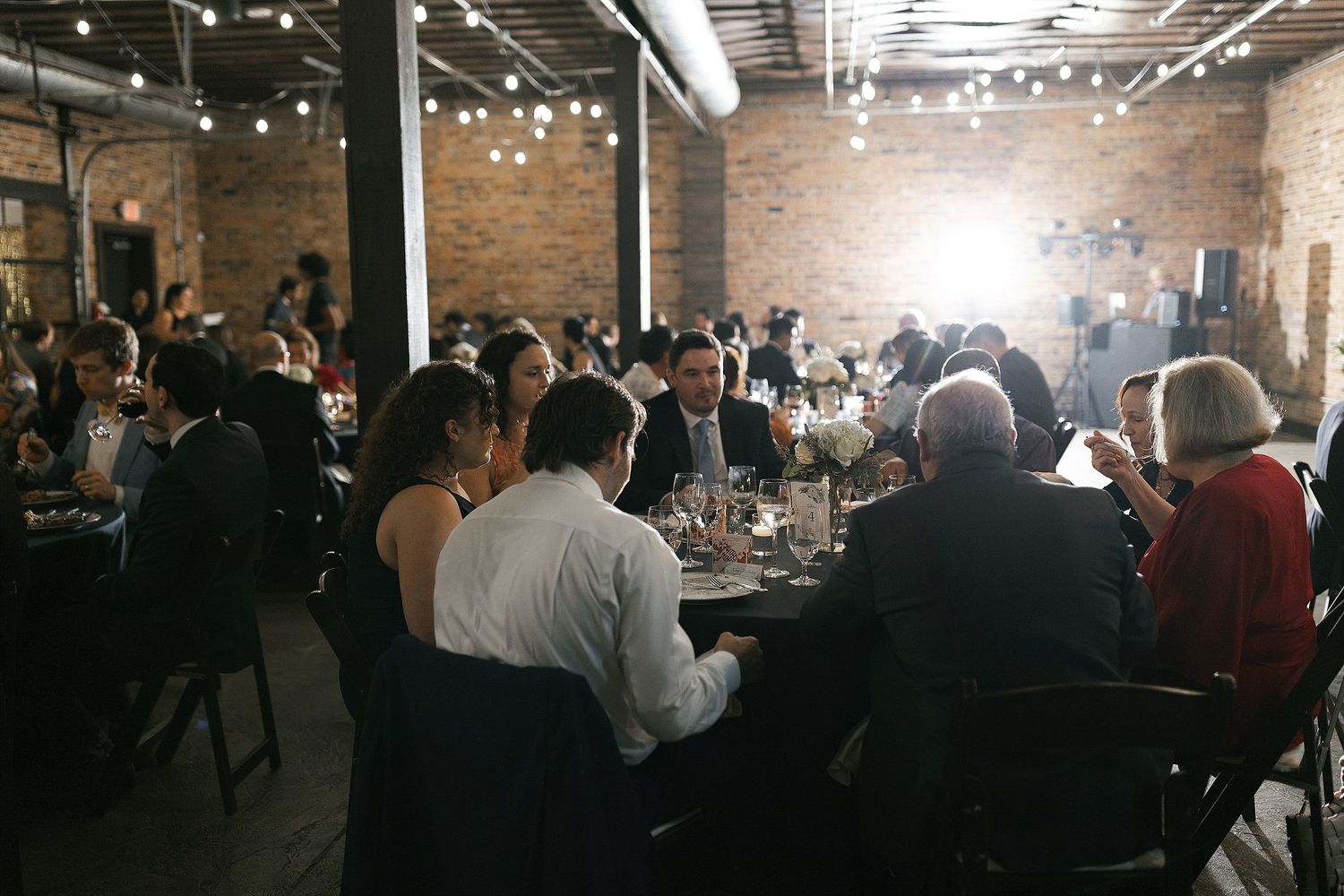 A candlelit reception dinner in an industrial brick venue with string lights hanging overhead and guests seated at long tables.