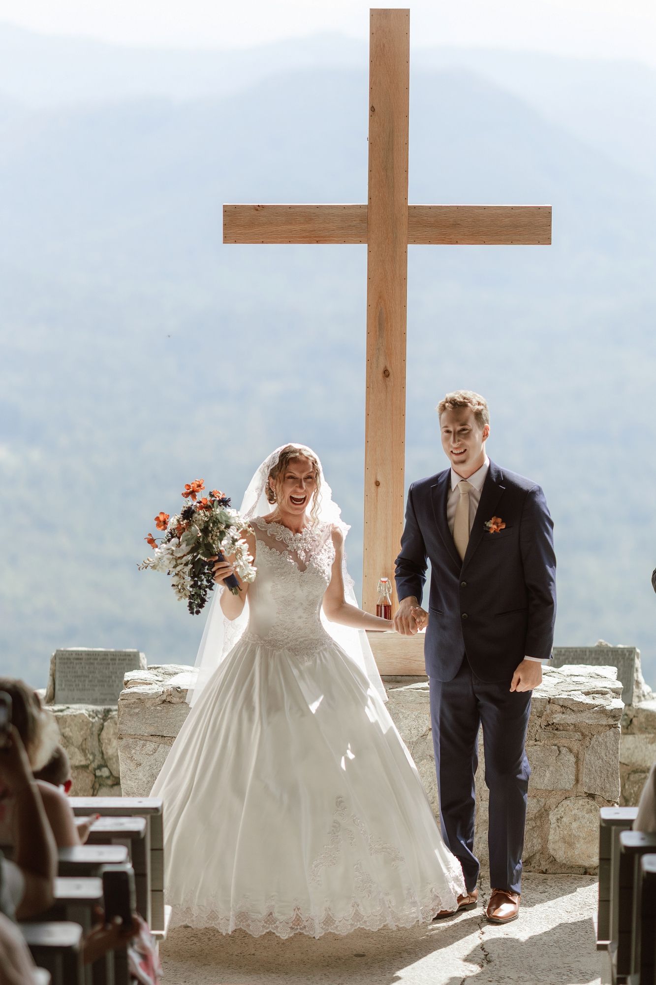 Joyful bride and groom walk down aisle before large wooden cross at mountain wedding ceremony.