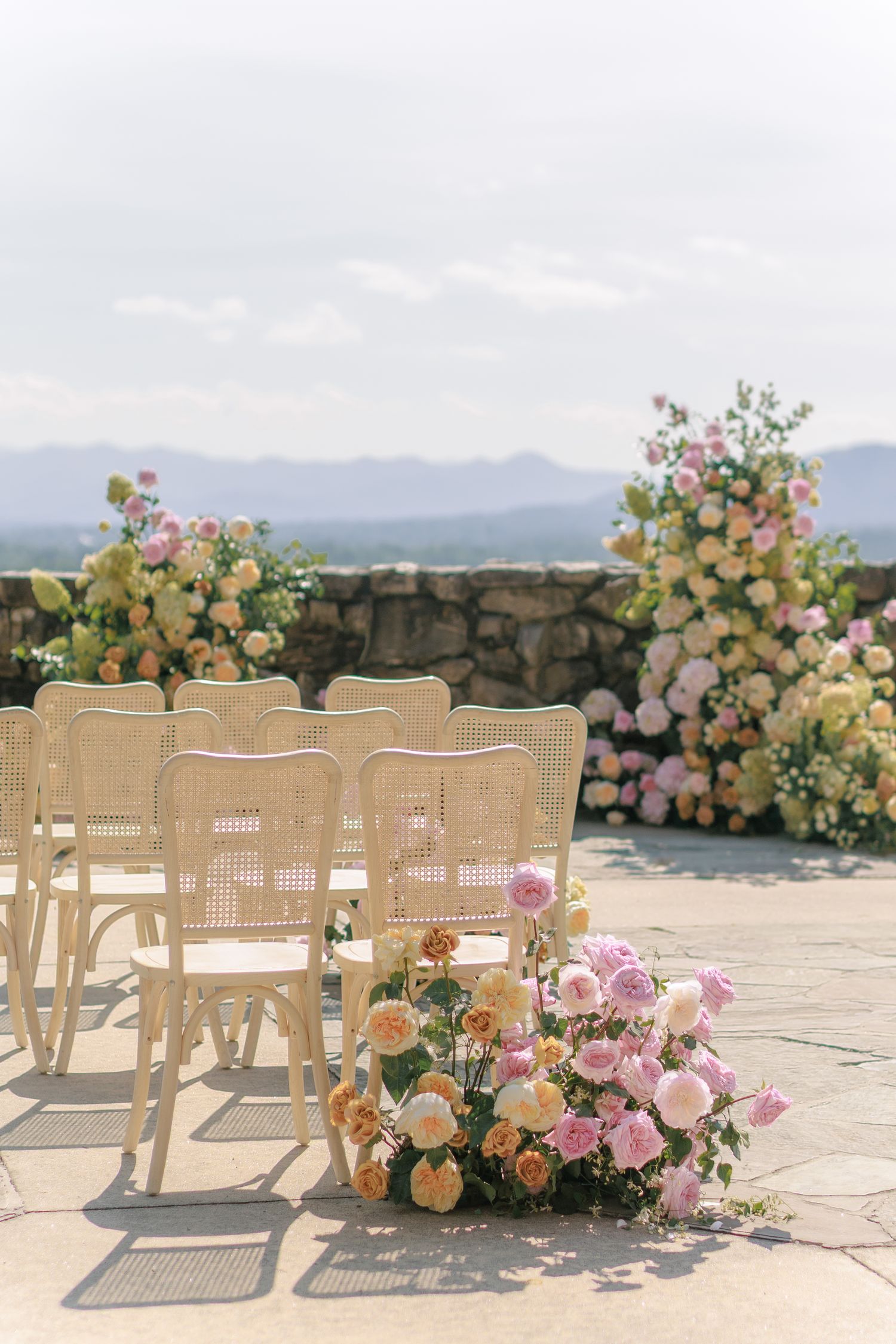 Elegant outdoor wedding ceremony with rattan chairs and pink, peach floral arrangements against a mountain backdrop.