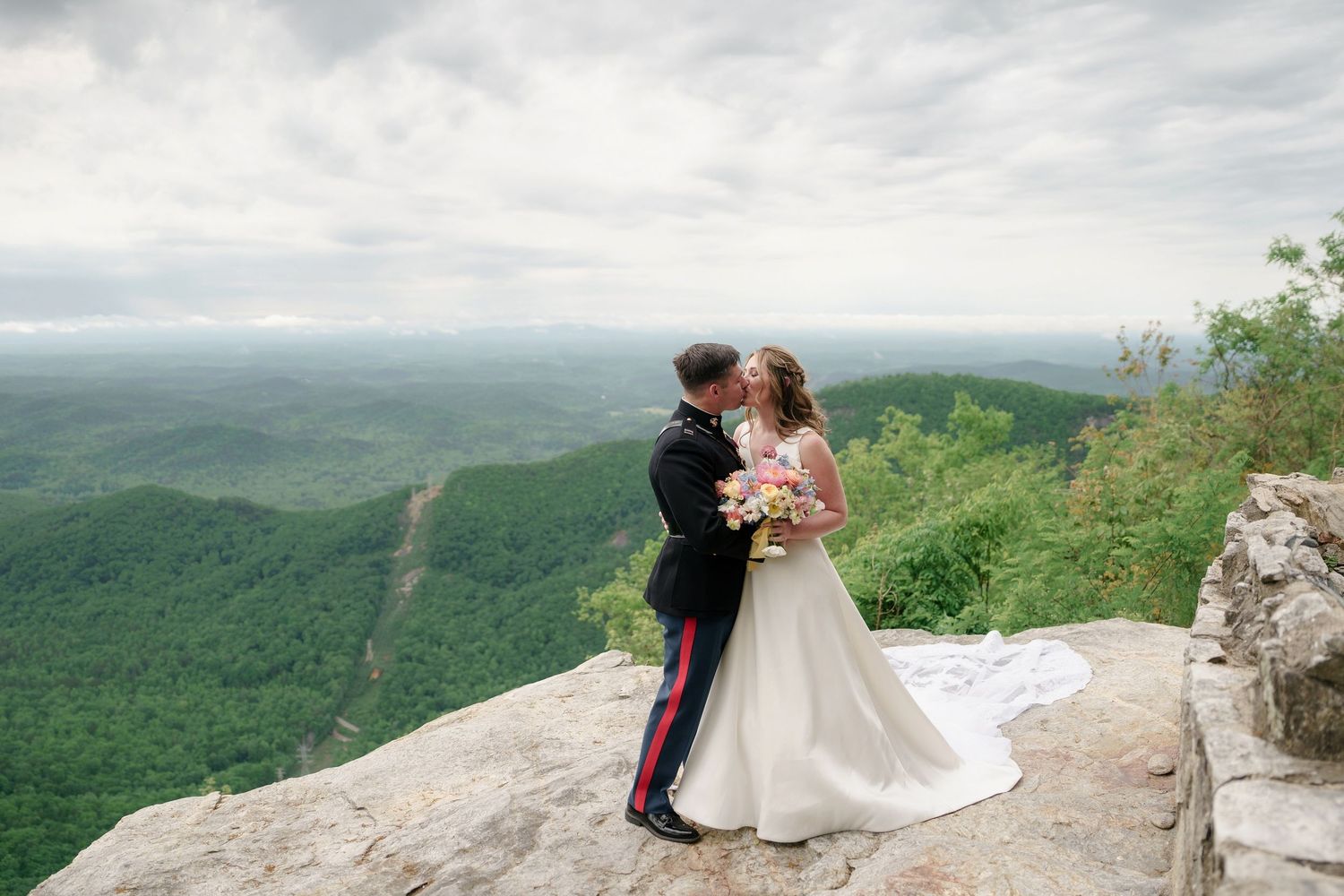 Military groom in dress uniform kisses bride on a dramatic rocky mountain cliff with lush valley below.