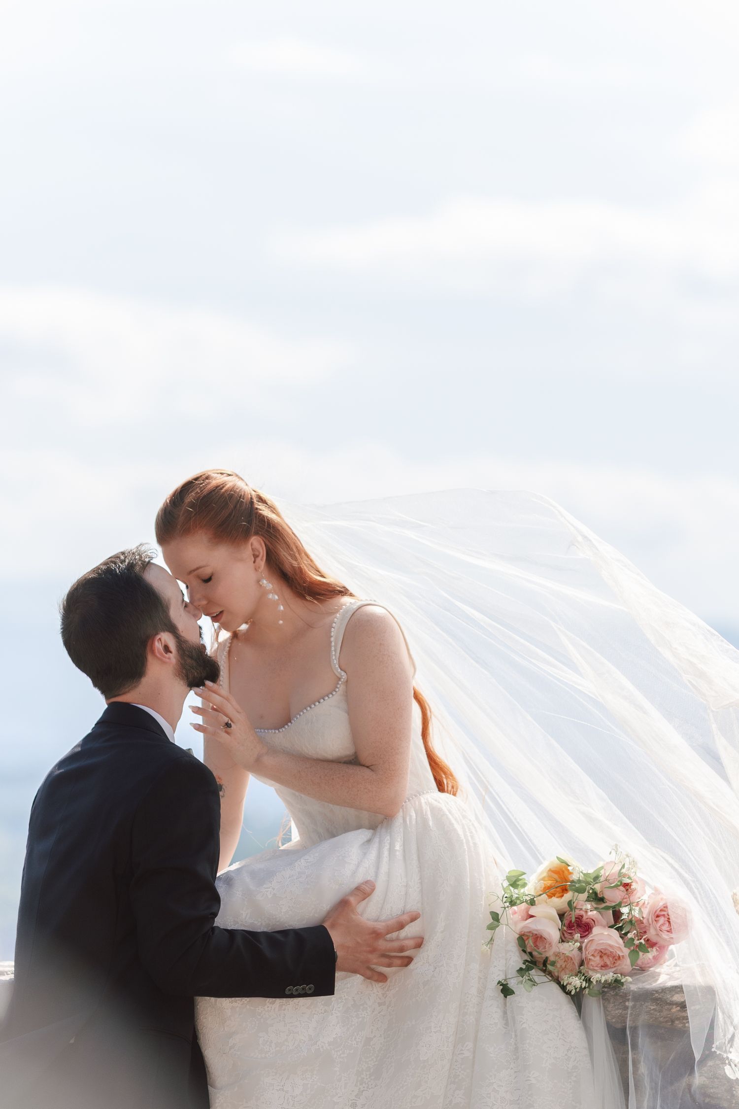 Bride and groom share romantic moment outdoors, her veil flowing in wind, holding pink bouquet against cloudy sky.