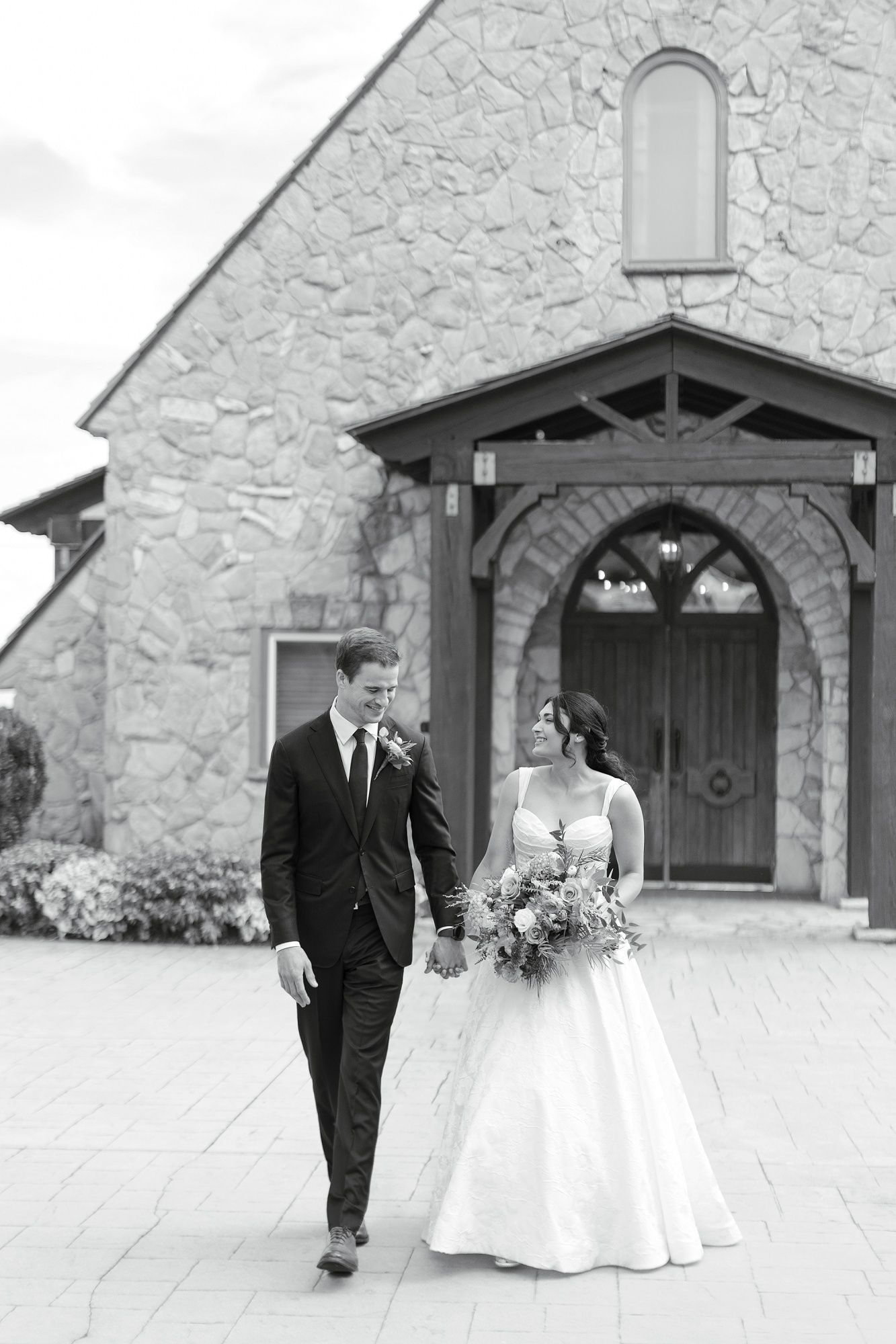 A bride and groom walk together outside a stone church building with arched doorways.