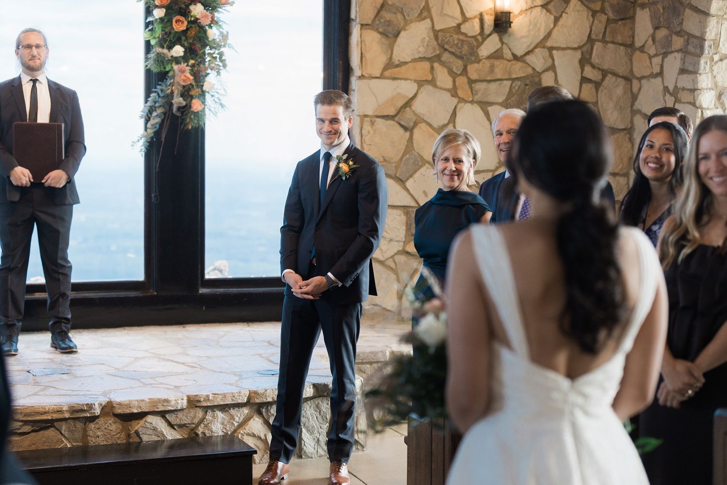 A wedding ceremony takes place in front of a rustic stone wall with guests watching the couple exchange vows.