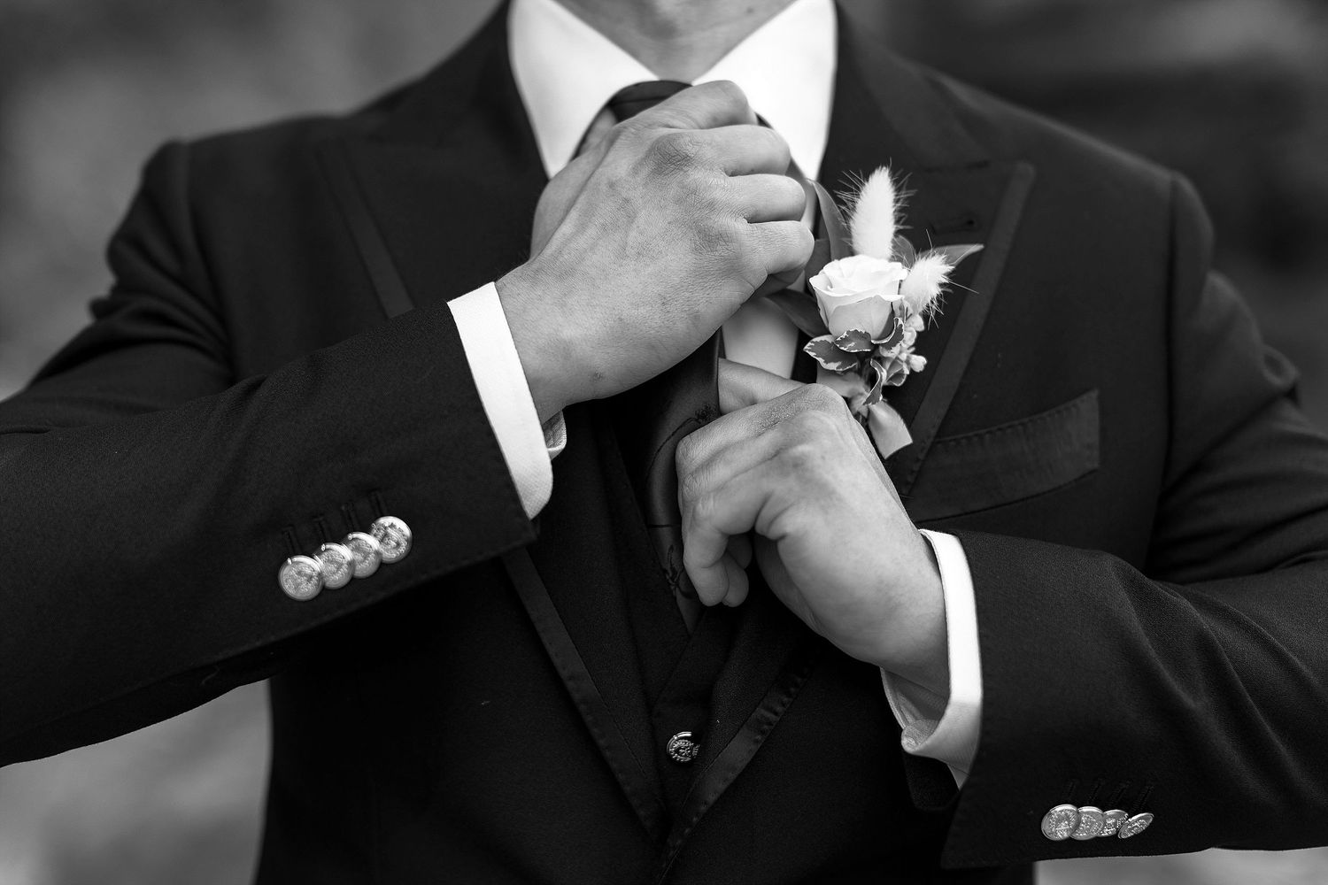 Black and white wedding photo showing hands adjusting tie and boutonniere on formal tuxedo suit.