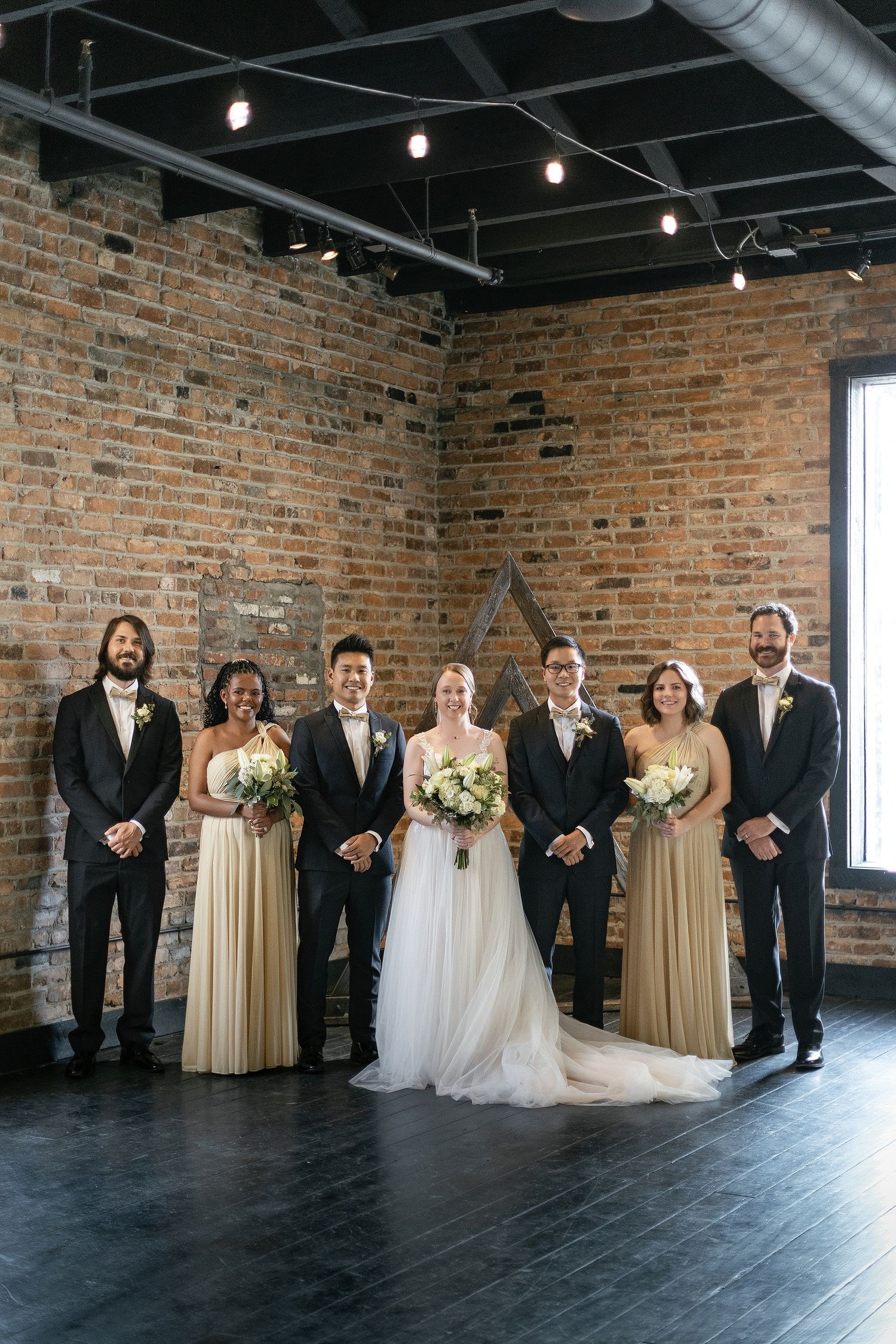 Wedding party poses in front of exposed brick wall in industrial venue with dark floors and hanging lights.