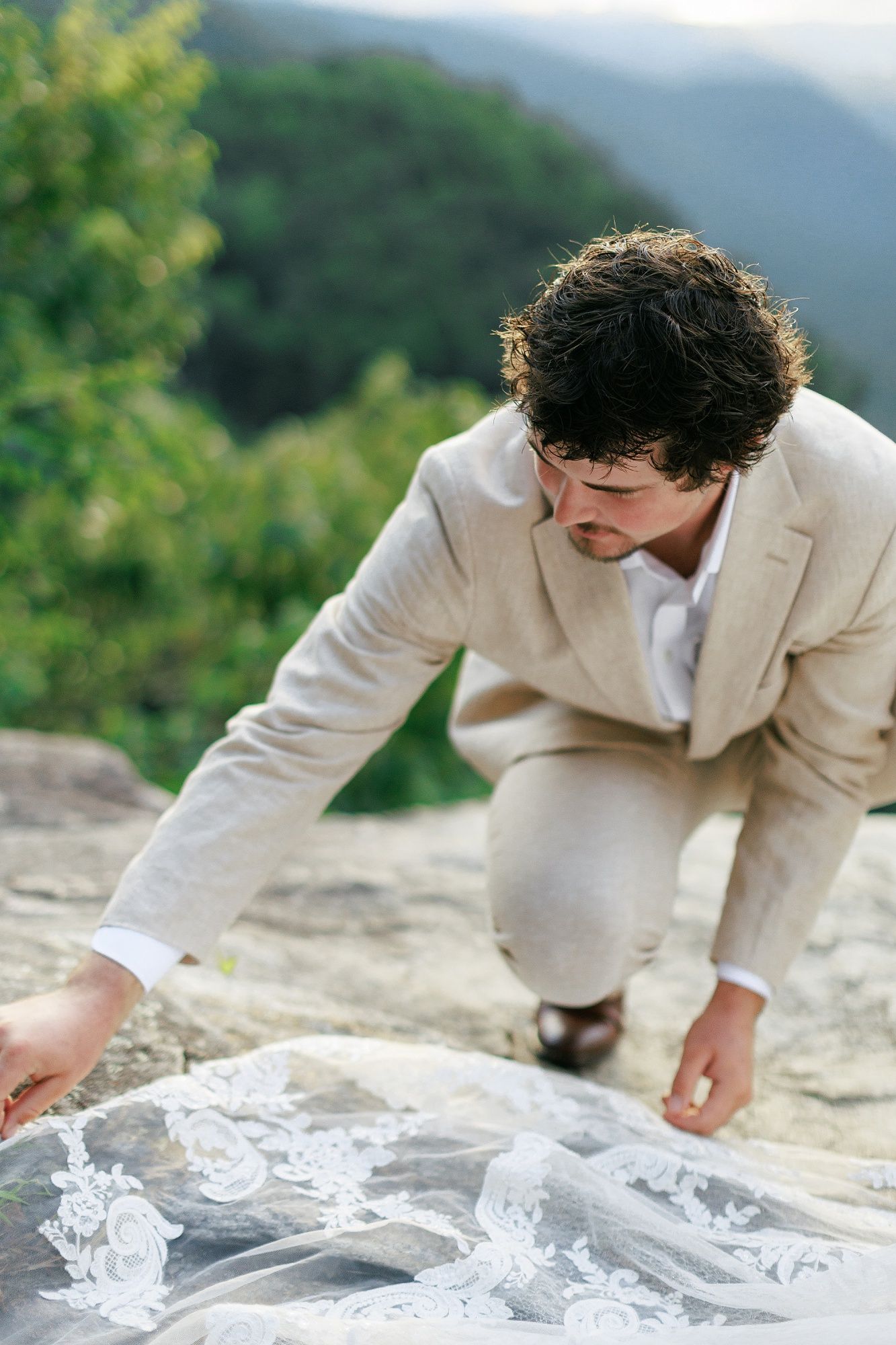 Someone wearing a light beige suit bends forward to unfold and arrange a delicate white fabric on a stone surface outdoors.