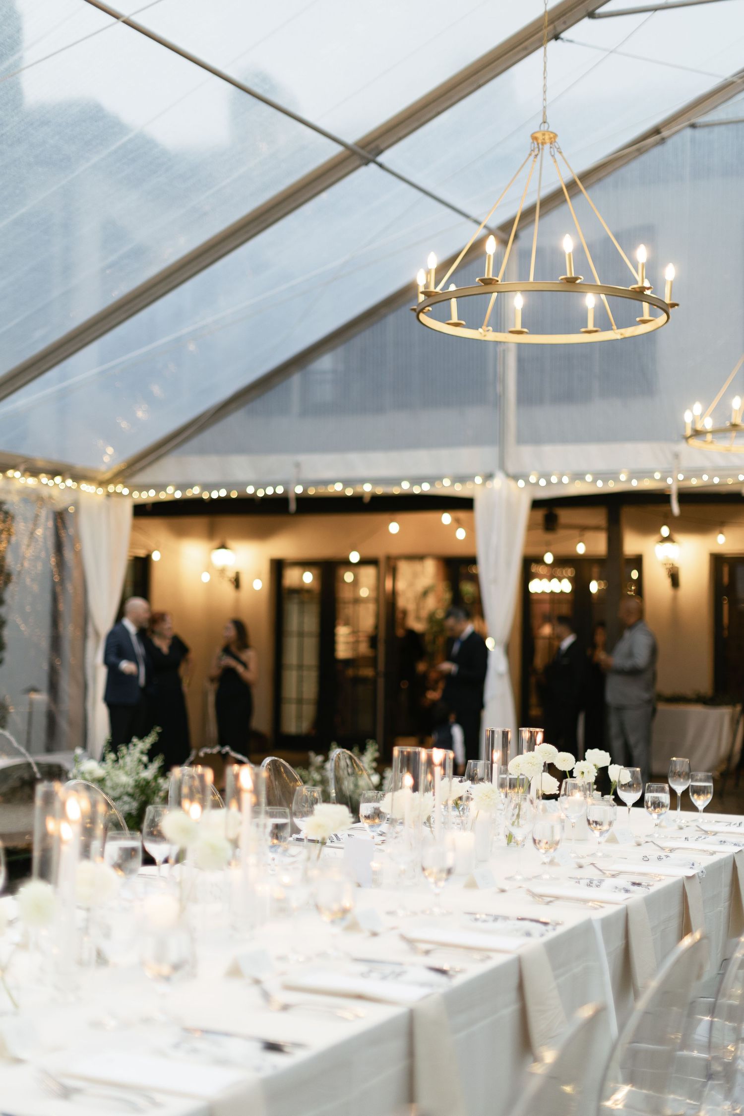 Elegant wedding reception table setting under a clear tent with circular chandeliers and soft lighting.
