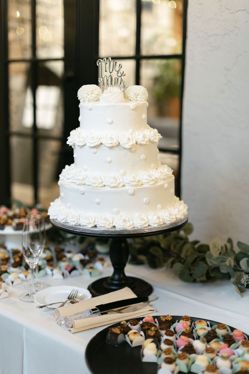 Classic three-tier white wedding cake with floral decorations and 'Mr & Mrs' topper on black stand.