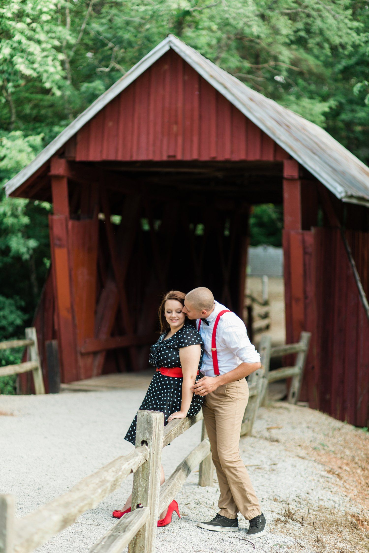 Couple poses playfully by red covered bridge during engagement session.