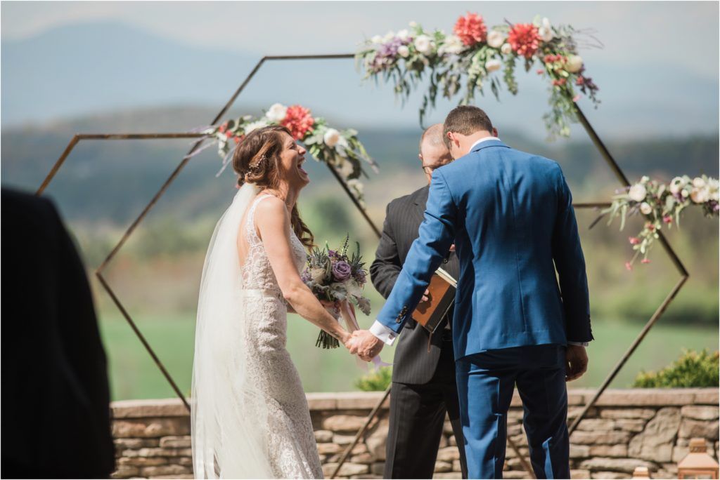 Couple exchanges vows under geometric arch decorated with pink and white flowers against mountain backdrop.
