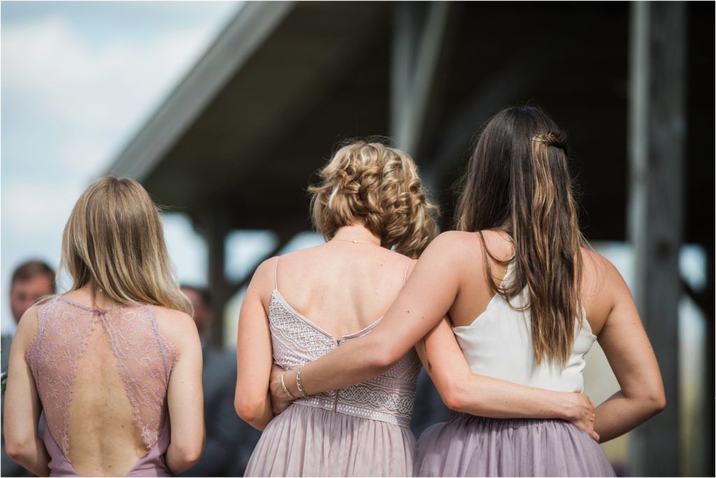 Three bridesmaids in pastel dresses standing with arms around each other's backs.
