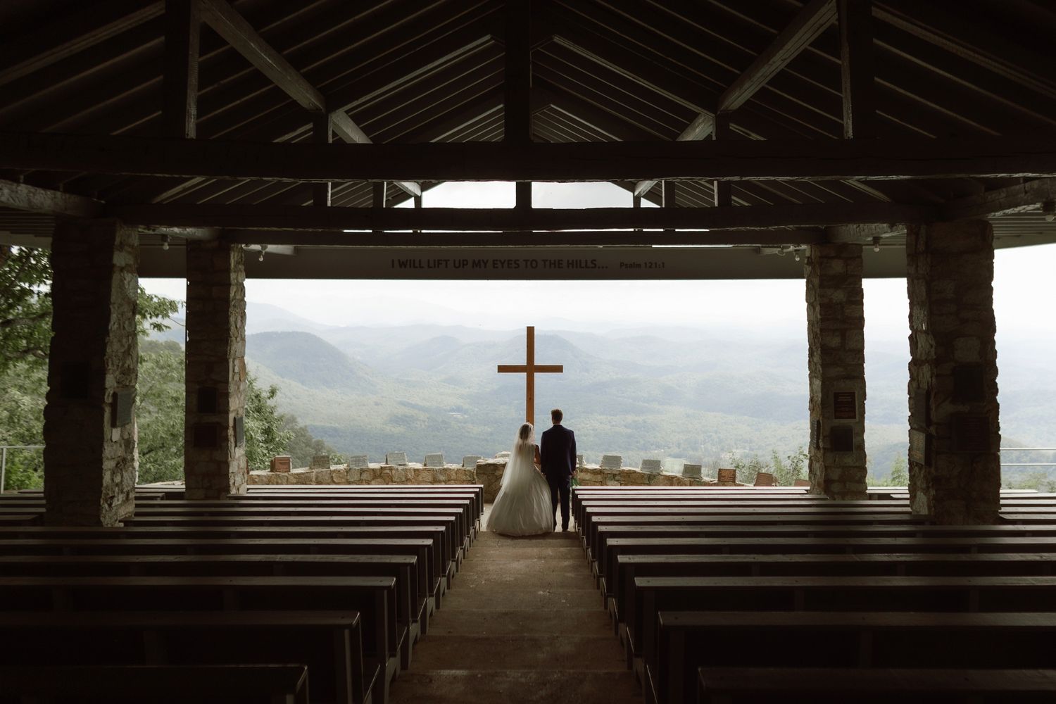 Couple standing before a wooden cross at an open-air mountain chapel with scenic views.