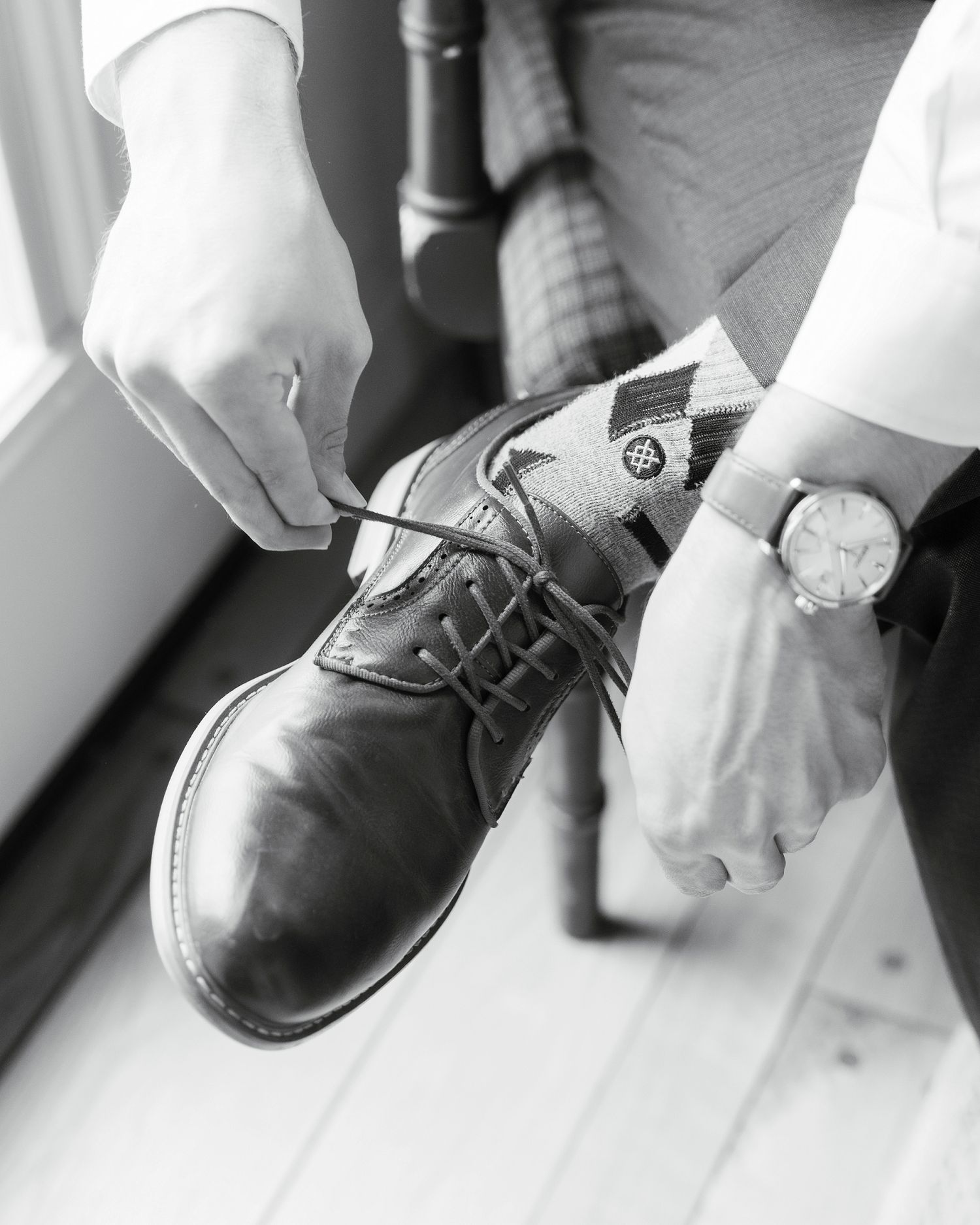 Groom tying dress shoe laces while wearing argyle socks and a watch, black and white wedding photo.