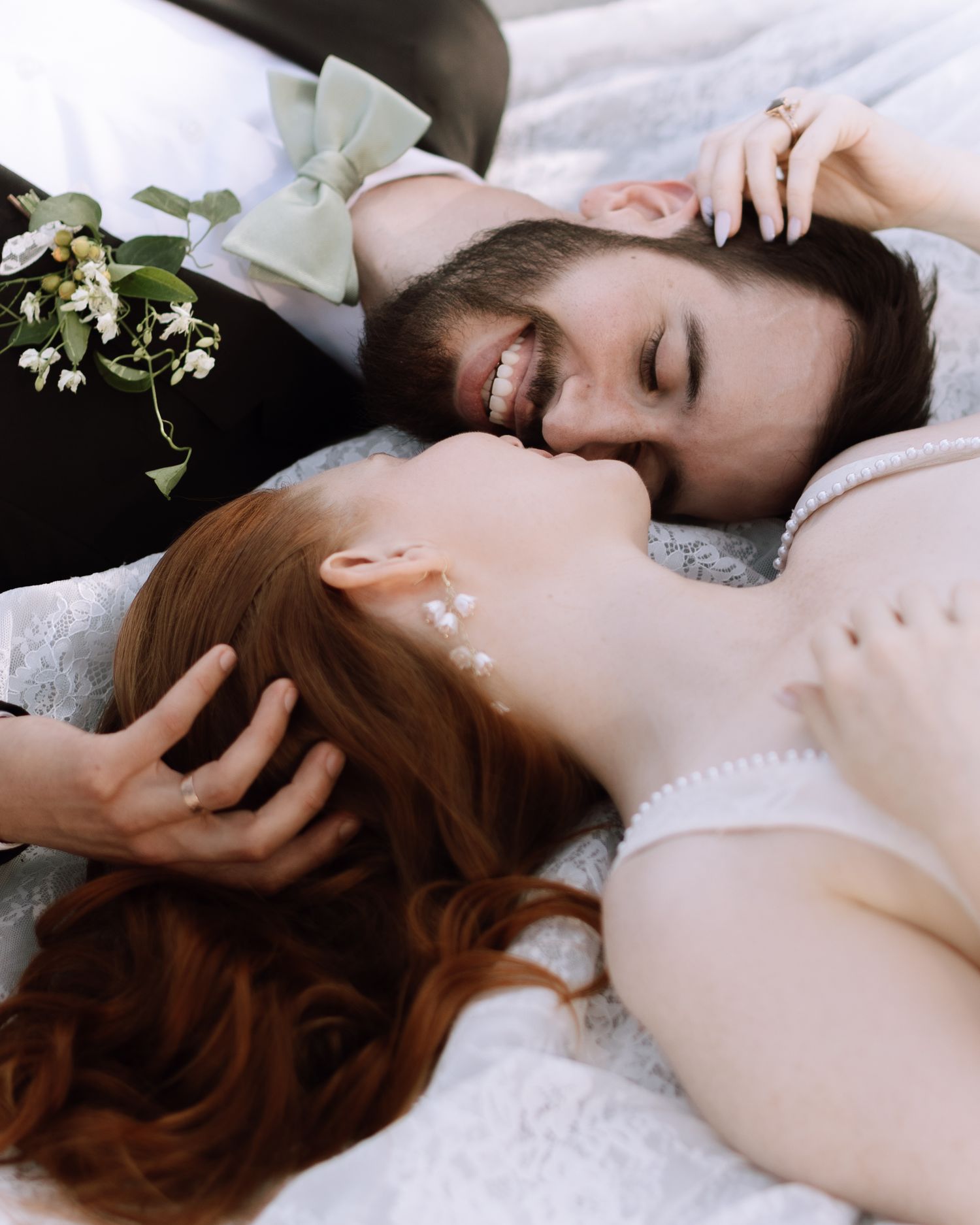 A smiling groom with beard gazes at his redhead bride as they lie together in wedding attire with floral accents.