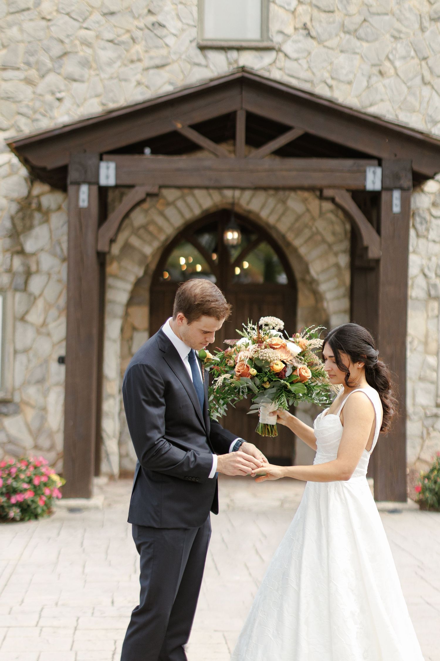Bride and groom share a tender moment outside a stone chapel, as she holds a lush orange and green wedding bouquet.