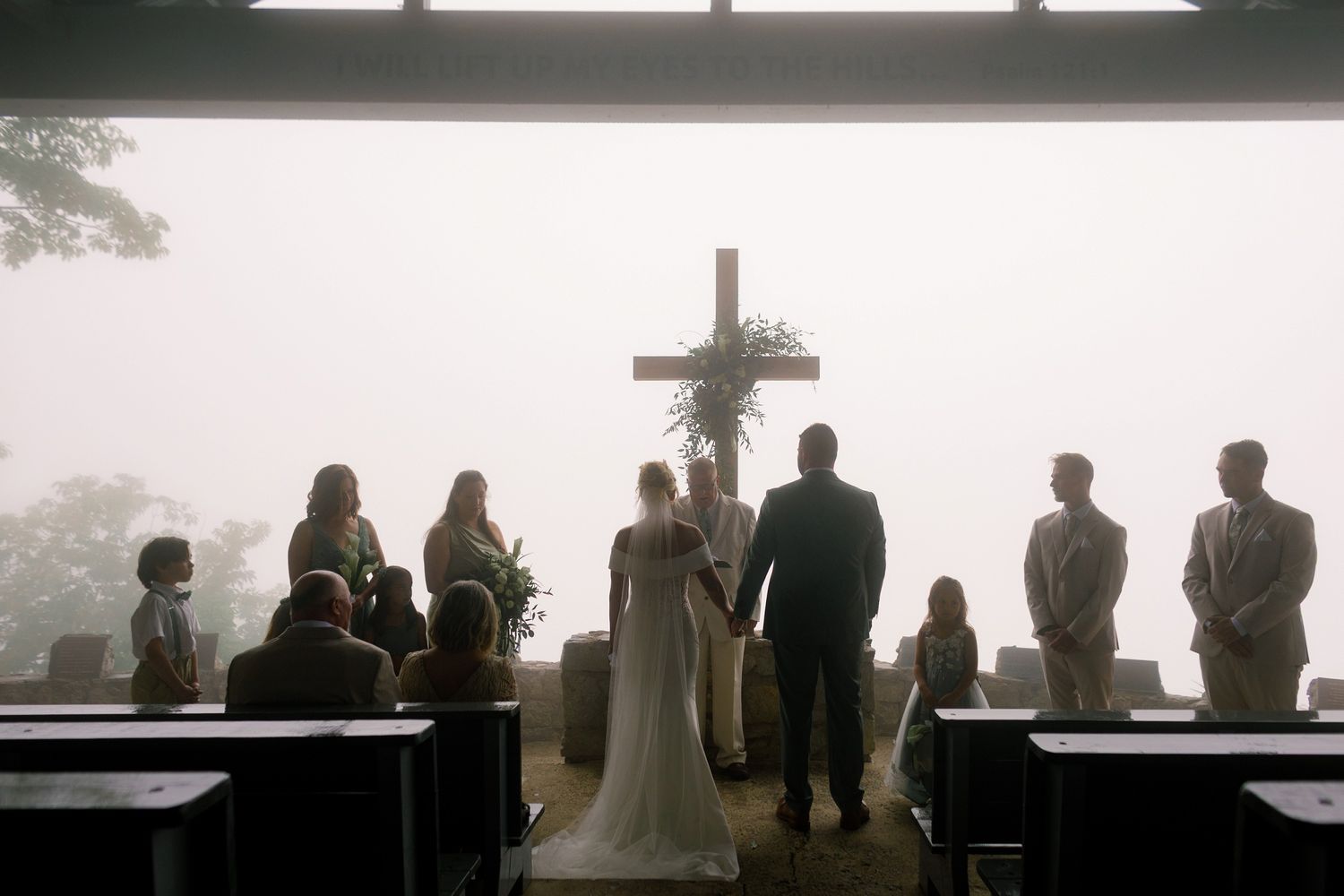 Silhouette of wedding ceremony at misty open-air chapel with wooden cross and mountain fog backdrop.