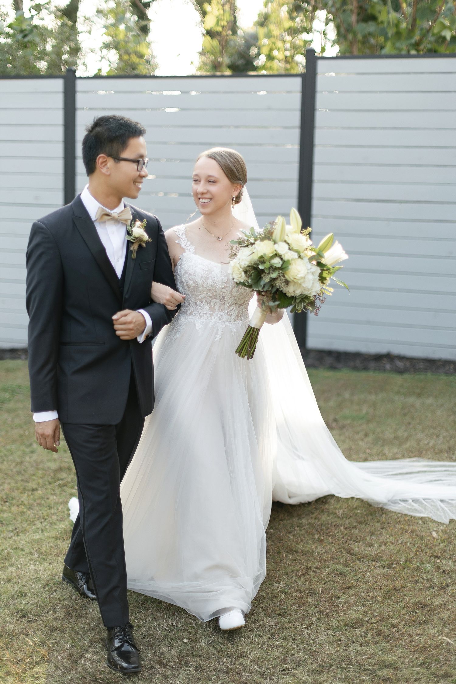 A couple in wedding attire walking together outdoors, bride holding white bouquet beside grey fence.