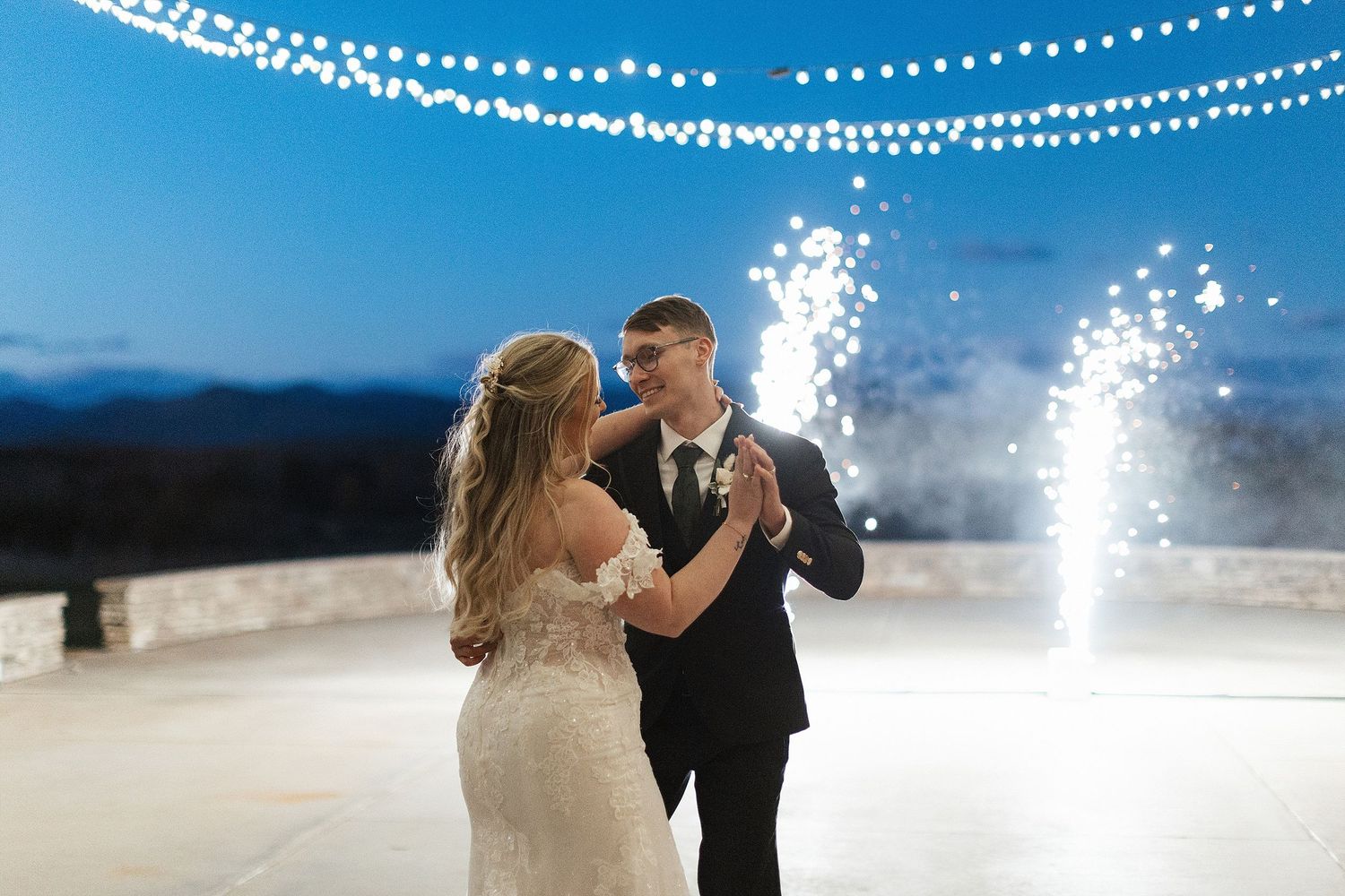 A couple shares their first dance outdoors at night with sparklers and string lights creating a magical atmosphere.