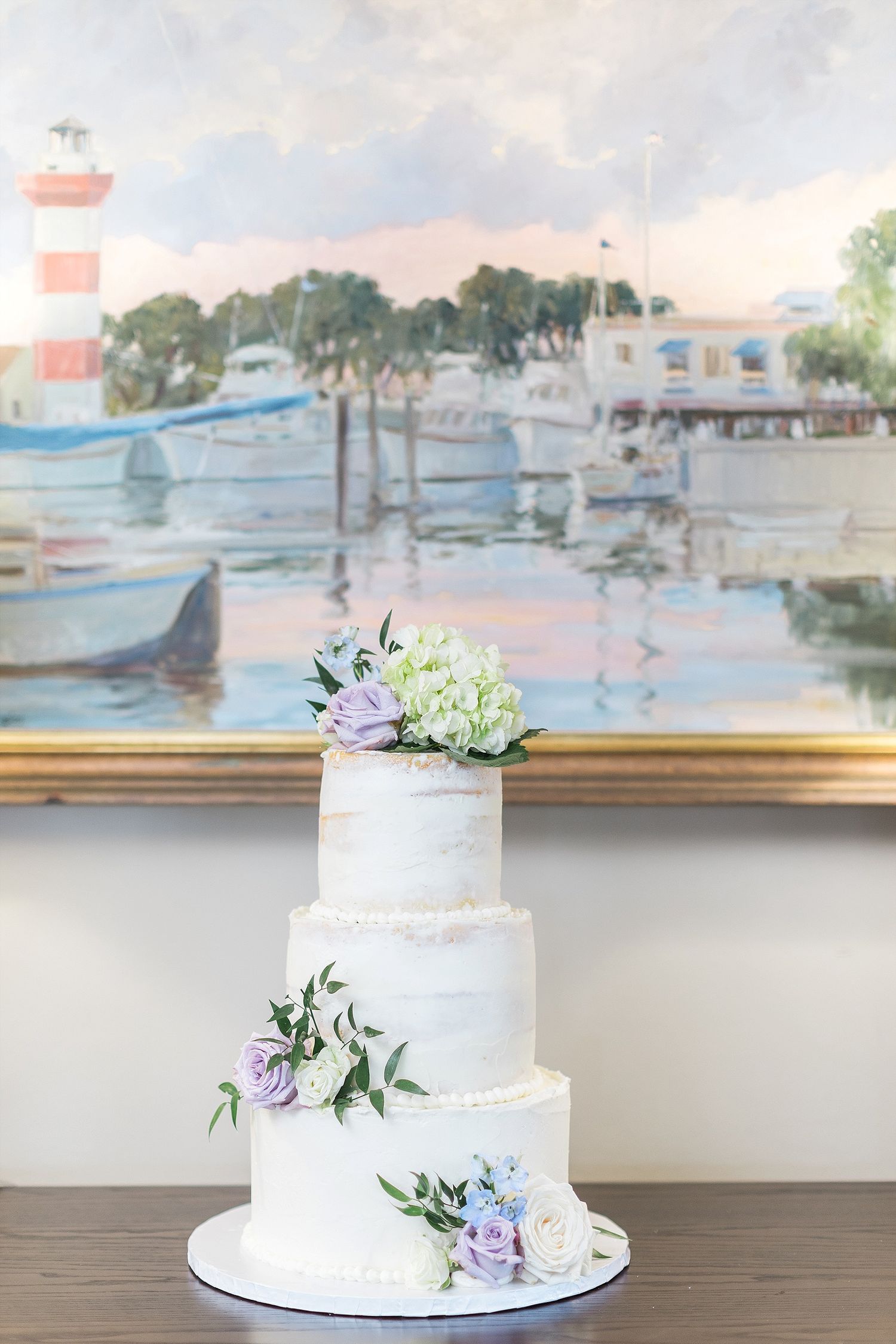 Three-tier white wedding cake decorated with pastel flowers against coastal artwork.