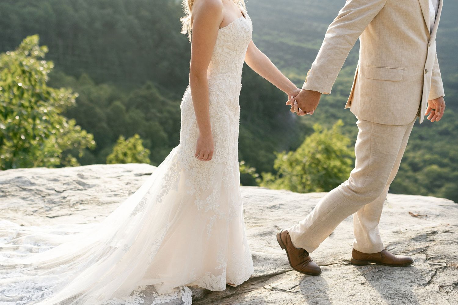 A couple wearing wedding attire stand together on a rocky mountain overlook against a scenic forest backdrop.
