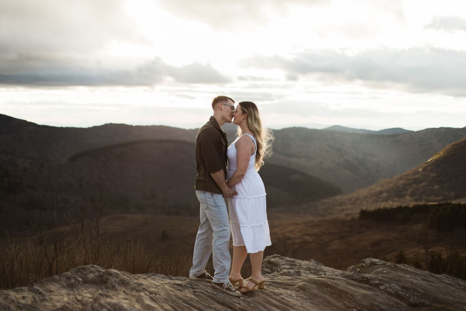 Two people embrace while standing on a mountain summit at sunset with dramatic cloudy skies and rolling hills in view.