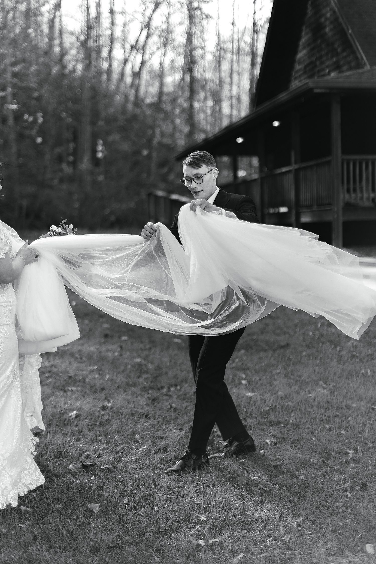 Black and white wedding photo showing flowing veil being held up near rustic cabin in wooded setting.