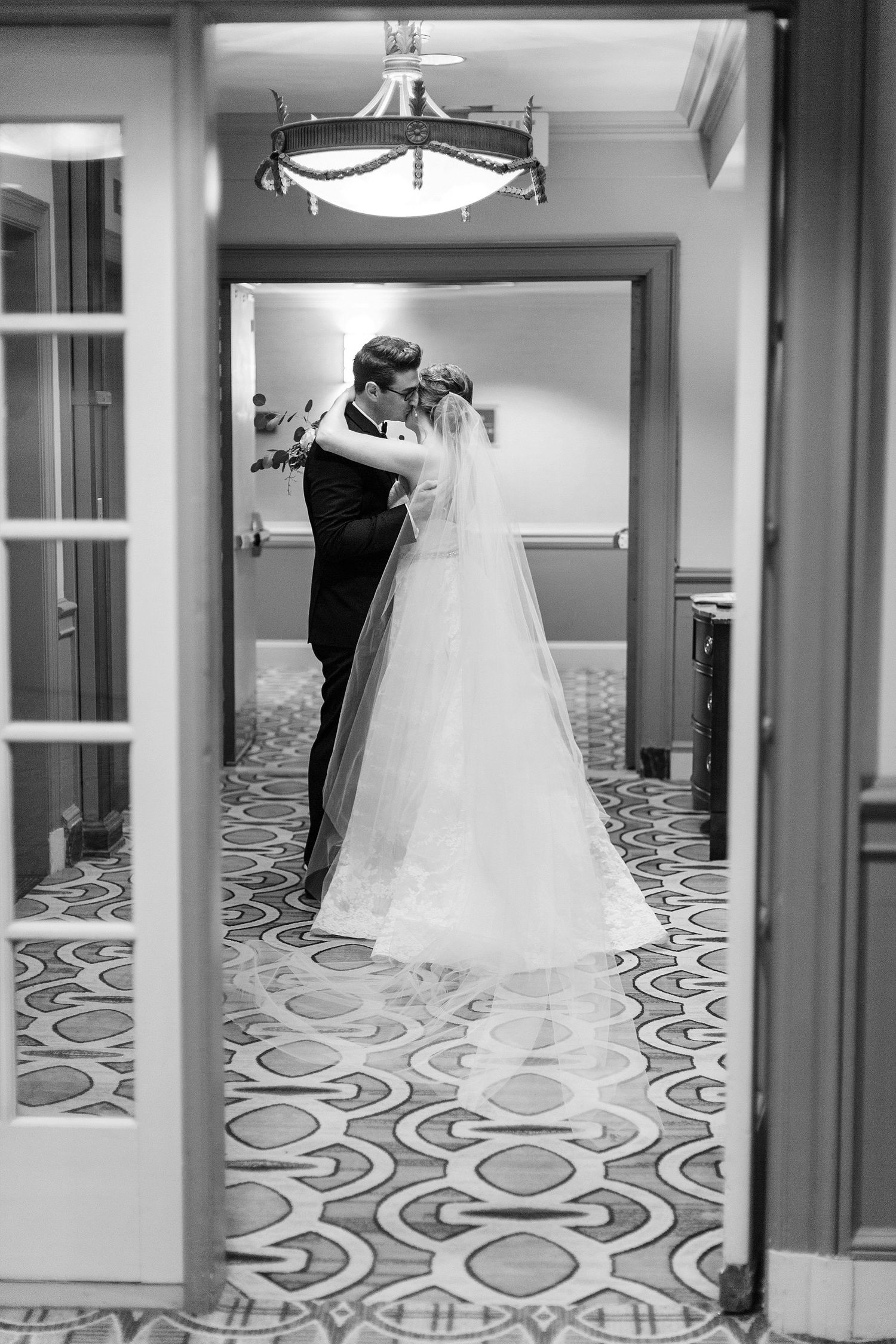 A black and white wedding photo showing a couple embracing in a hallway with decorative patterned carpet.