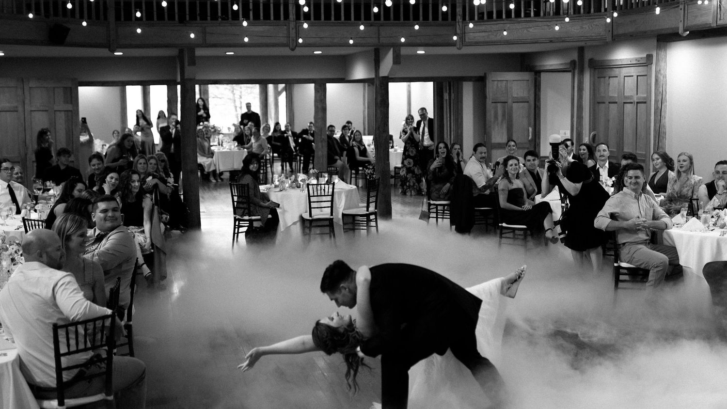 Wedding guests observe dancers performing on a low-lit dance floor during an indoor reception celebration.
