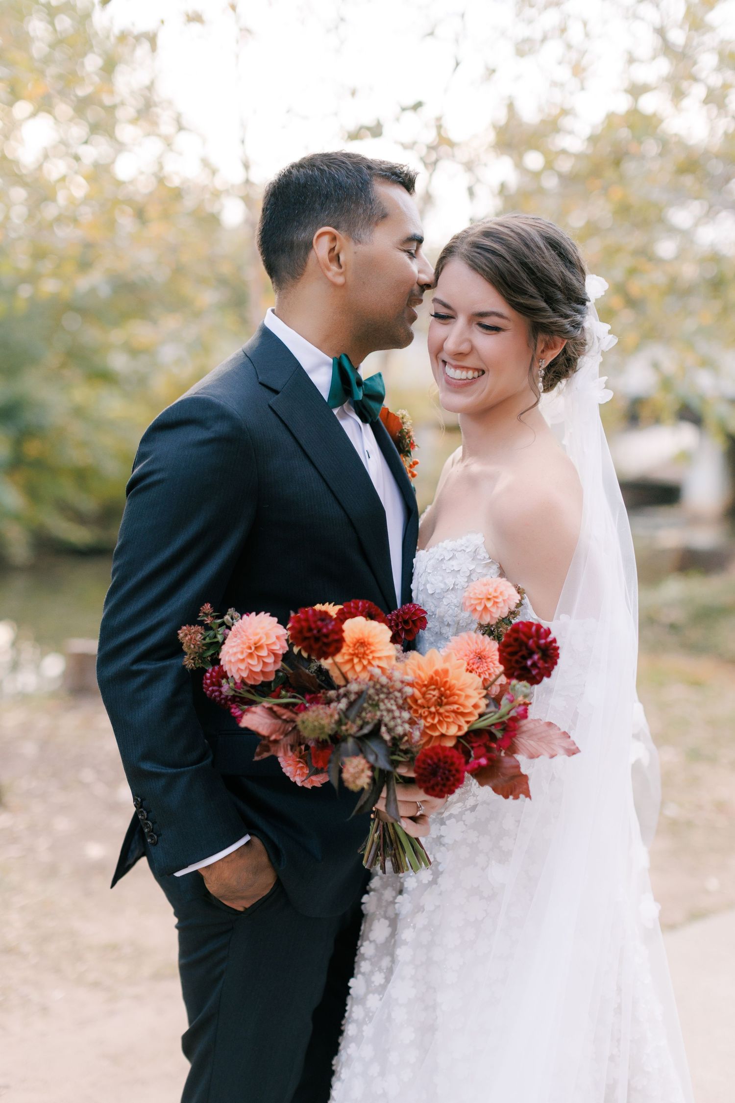 Groom in navy suit kisses bride wearing white floral gown holding autumn dahlia bouquet outside the Huguenot Mill and Loft in Greenville, SC