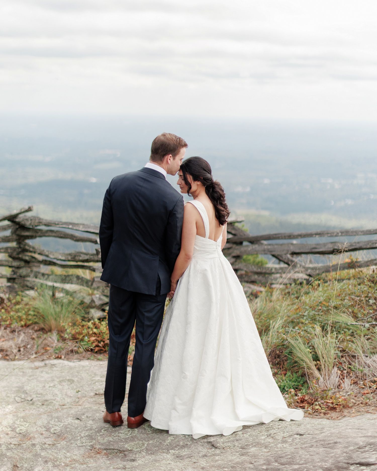 A couple in wedding attire share an intimate moment on a scenic mountain overlook with a misty valley below.