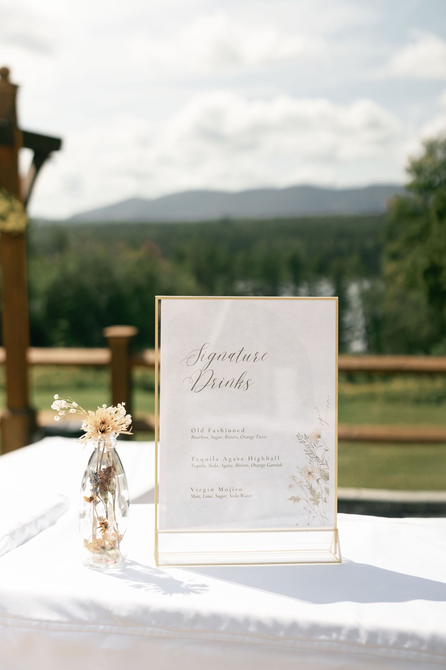 Elegant wedding signature drinks menu in gold frame on white table with small floral arrangement and mountain backdrop.