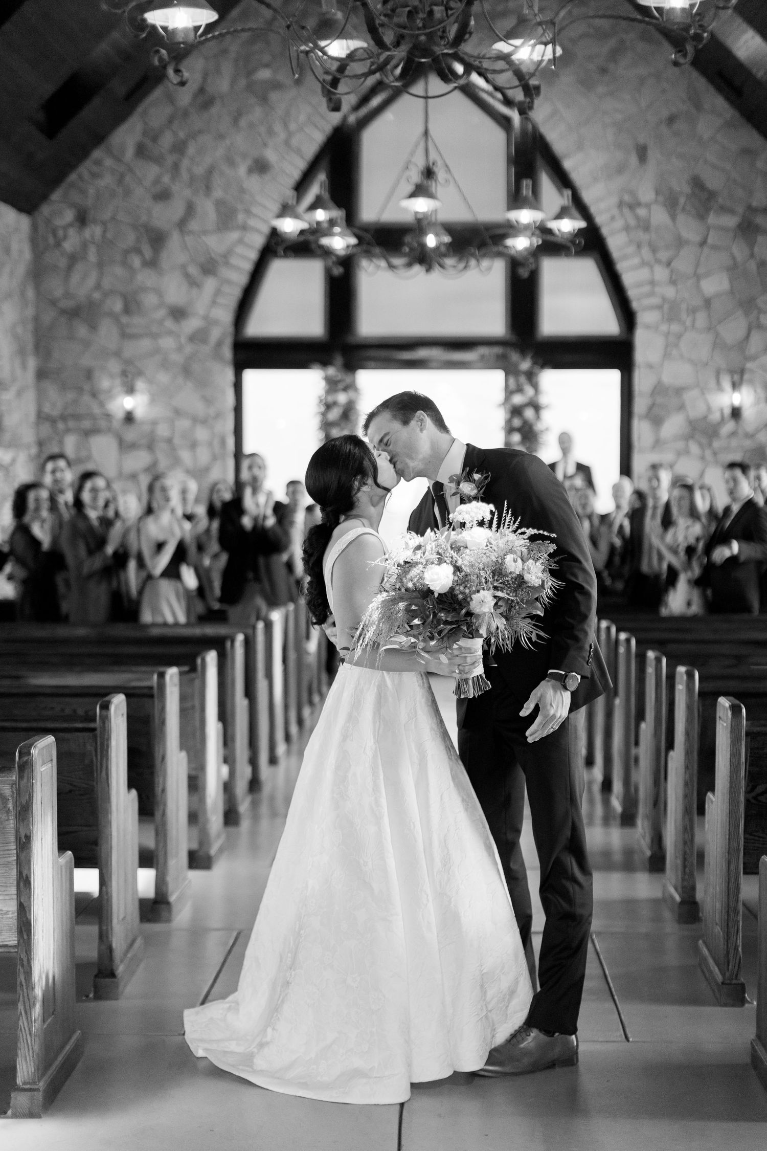 Black and white photo of a bride and groom sharing their first kiss at the altar in a rustic stone chapel.