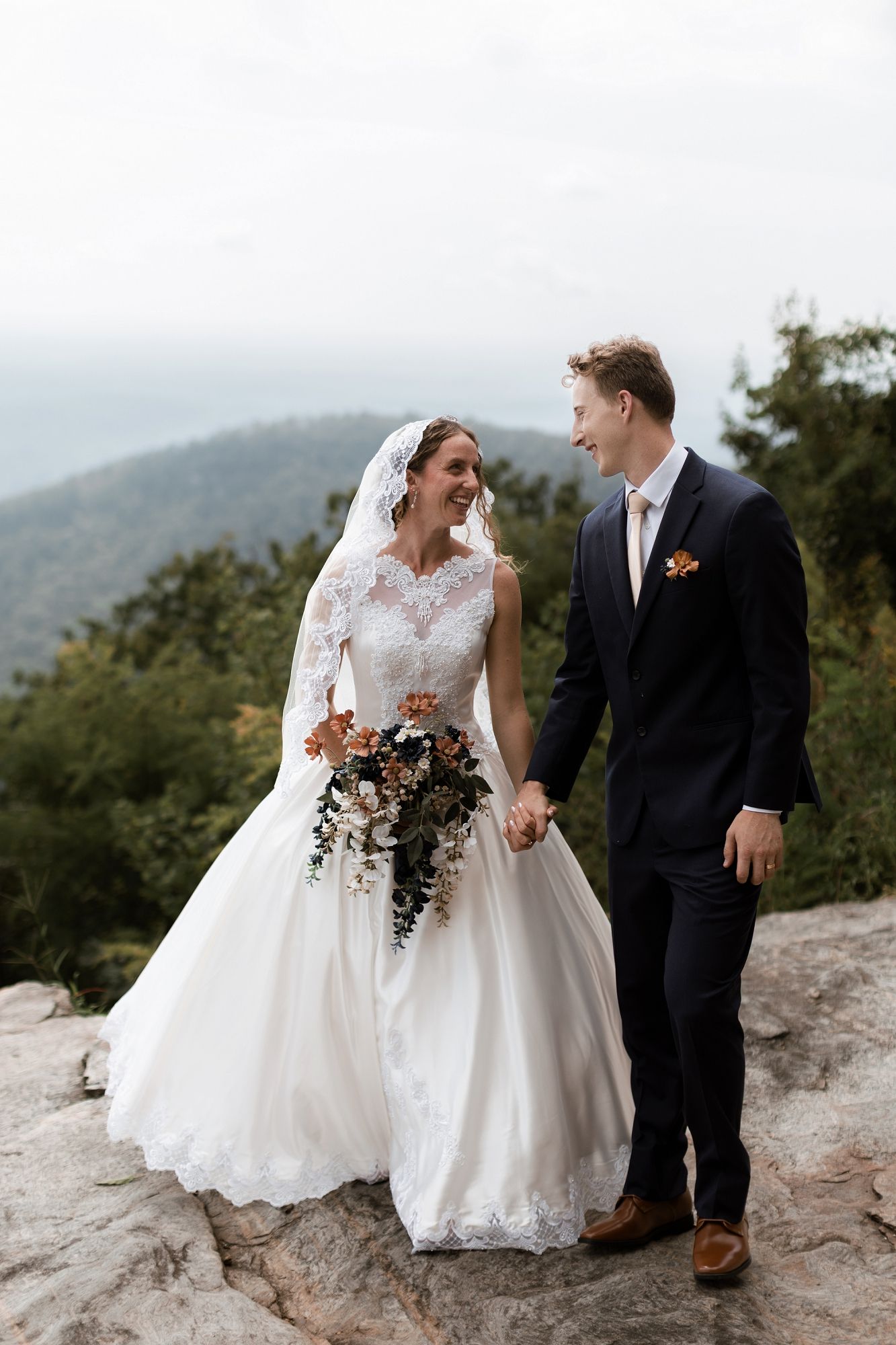 Laughing bride in lace gown and groom in navy suit hold hands on rocky mountain overlook.