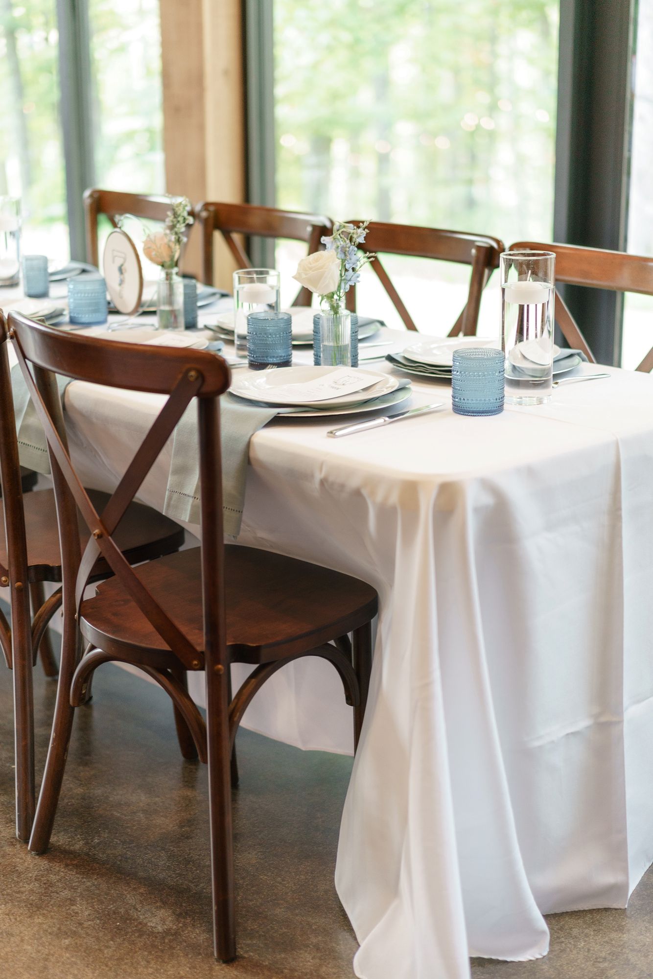 Elegant wedding reception table with white linen, wooden cross-back chairs, blue candles, and simple floral centerpieces.