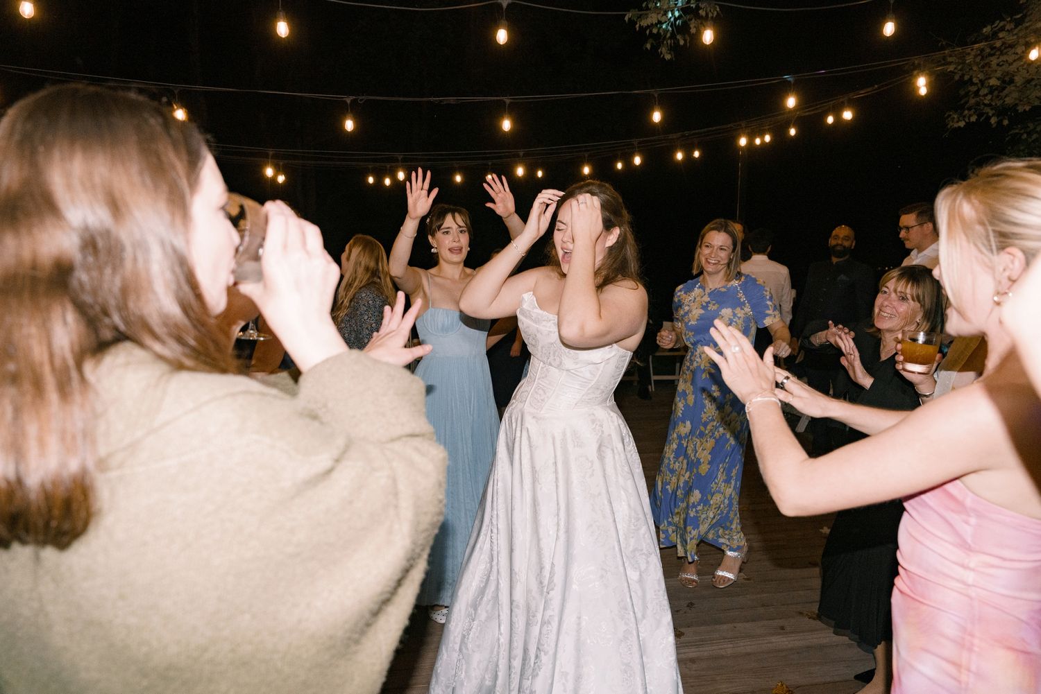 Bride in white gown dancing joyfully with guests under string lights at an outdoor wedding reception.