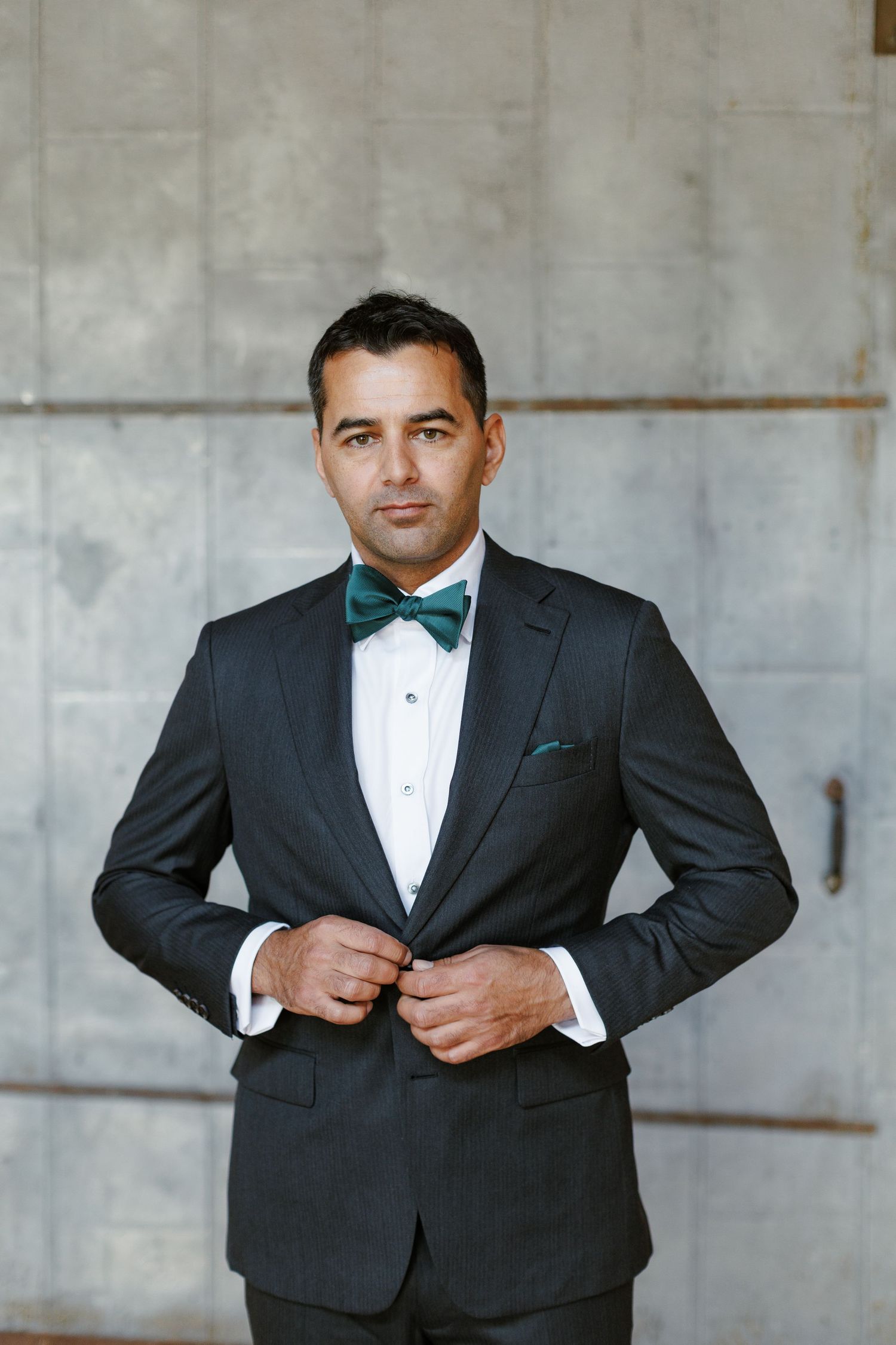 Man in black tuxedo with green bow tie and pocket square buttoning jacket against concrete wall.