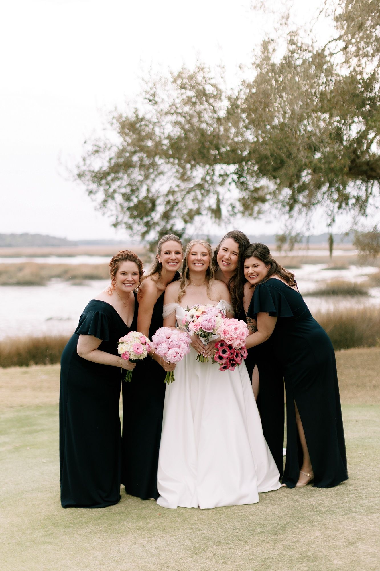 Bridal party laughs together on a scenic lawn with water and trees in the background.