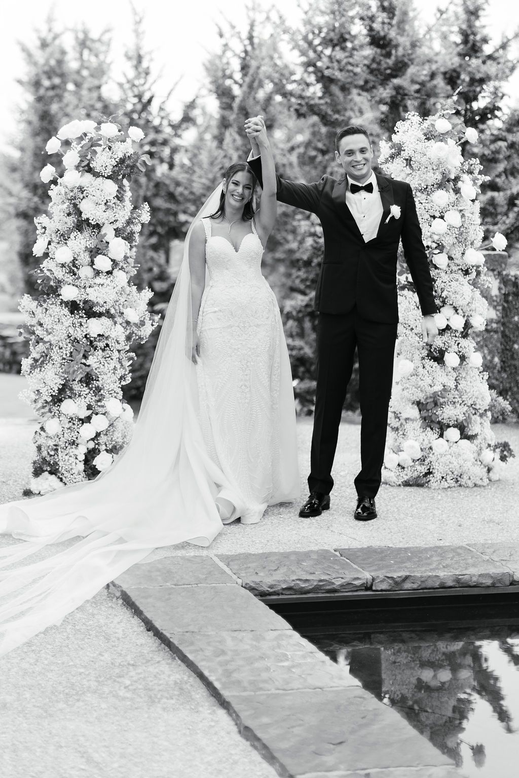A wedding couple celebrates under a floral arch by a reflecting pool in a black and white photograph.