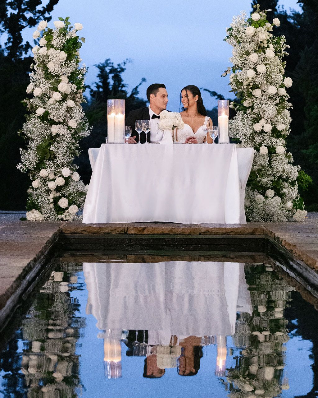 Elegant wedding sweetheart table setup with white floral arrangements reflected in pool at twilight.